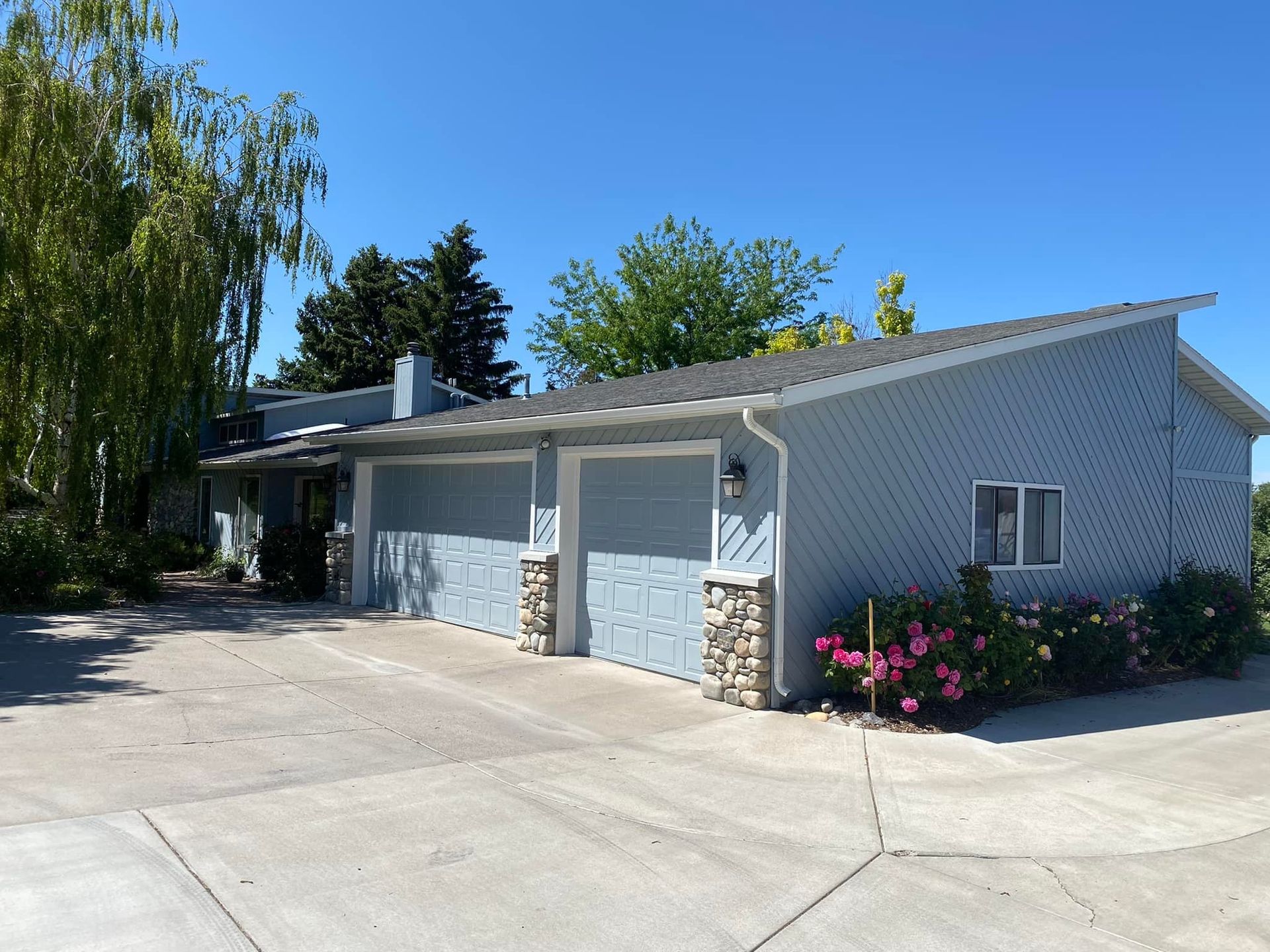 Blue house with two garage doors and stone accents, concrete driveway, and flowering bushes.
