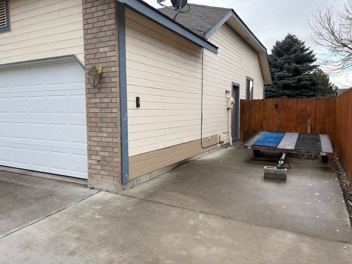 Garage exterior with concrete driveway, wooden fence, picnic table, tan siding, brick chimney, and overcast sky.