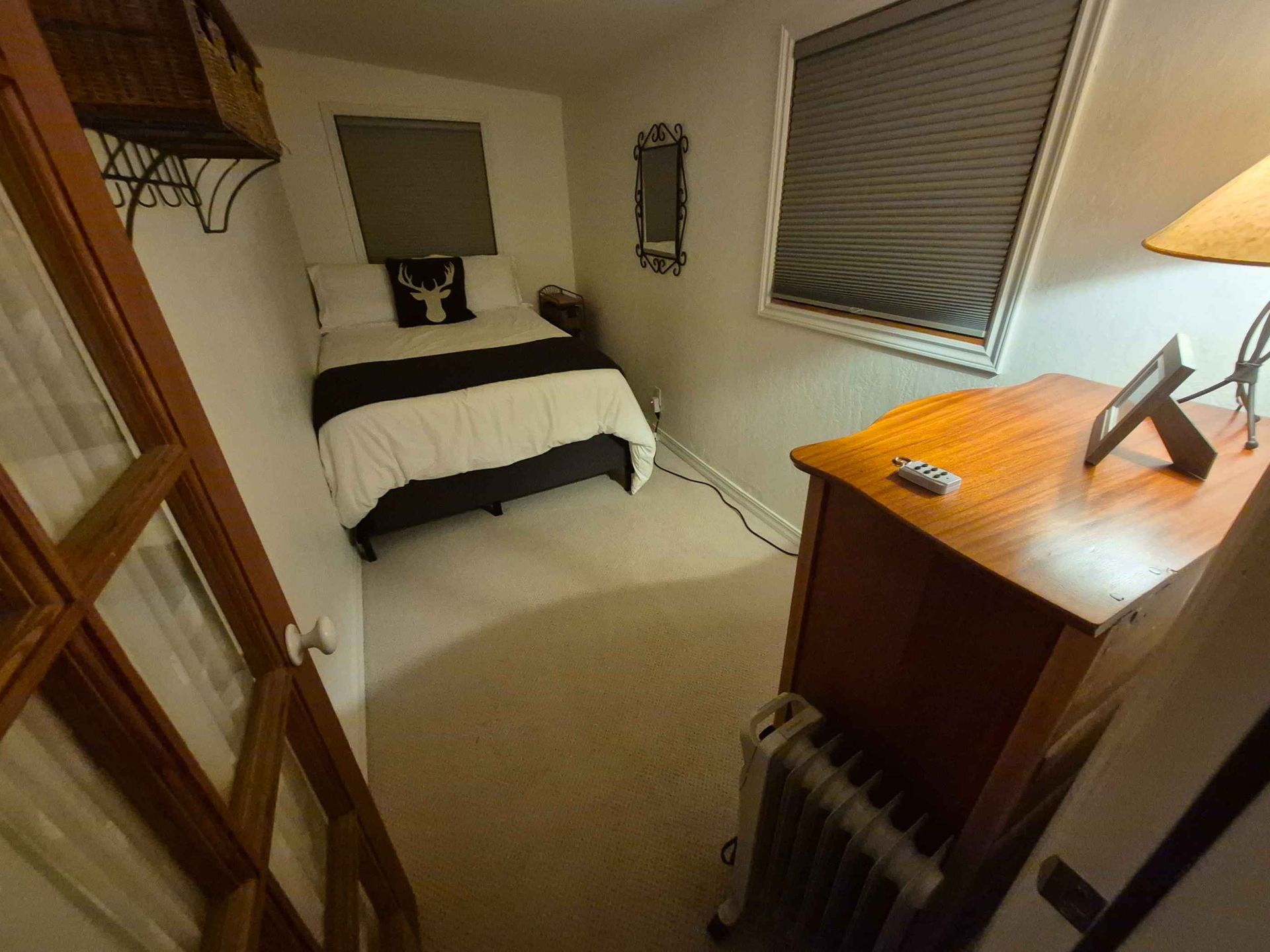 Small bedroom with a single bed, wooden dresser, window blinds, and a desk lamp.