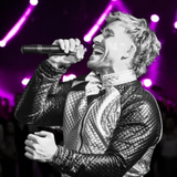 Cantante actuando frente a un micrófono bajo luces de escenario moradas, vistiendo una chaqueta acolchada negra.
