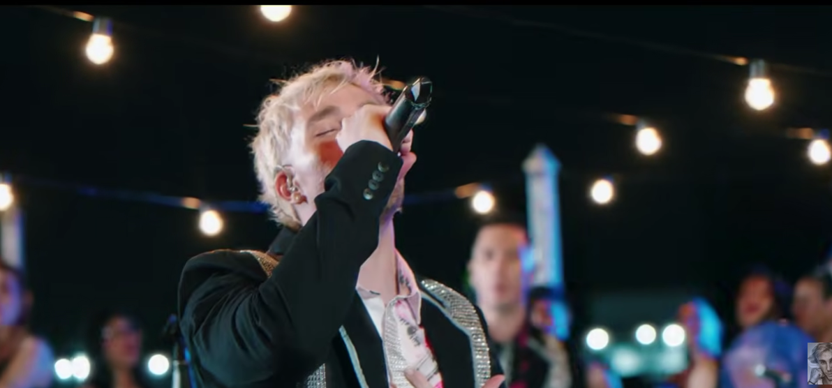 Cantante sosteniendo un micrófono en el escenario bajo luces de guirnalda, con personas borrosas al fondo.