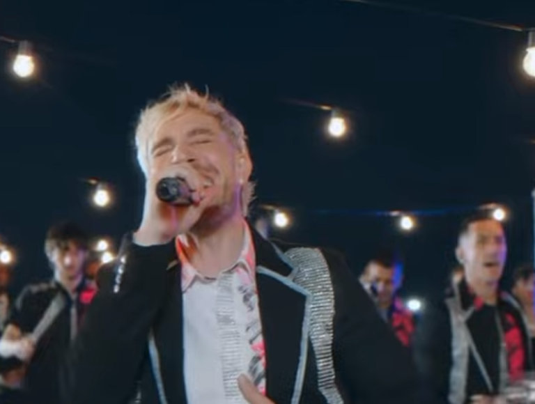 Cantante actuando con micrófono en un escenario iluminado, vistiendo una chaqueta negra con ribetes plateados y sonriendo.