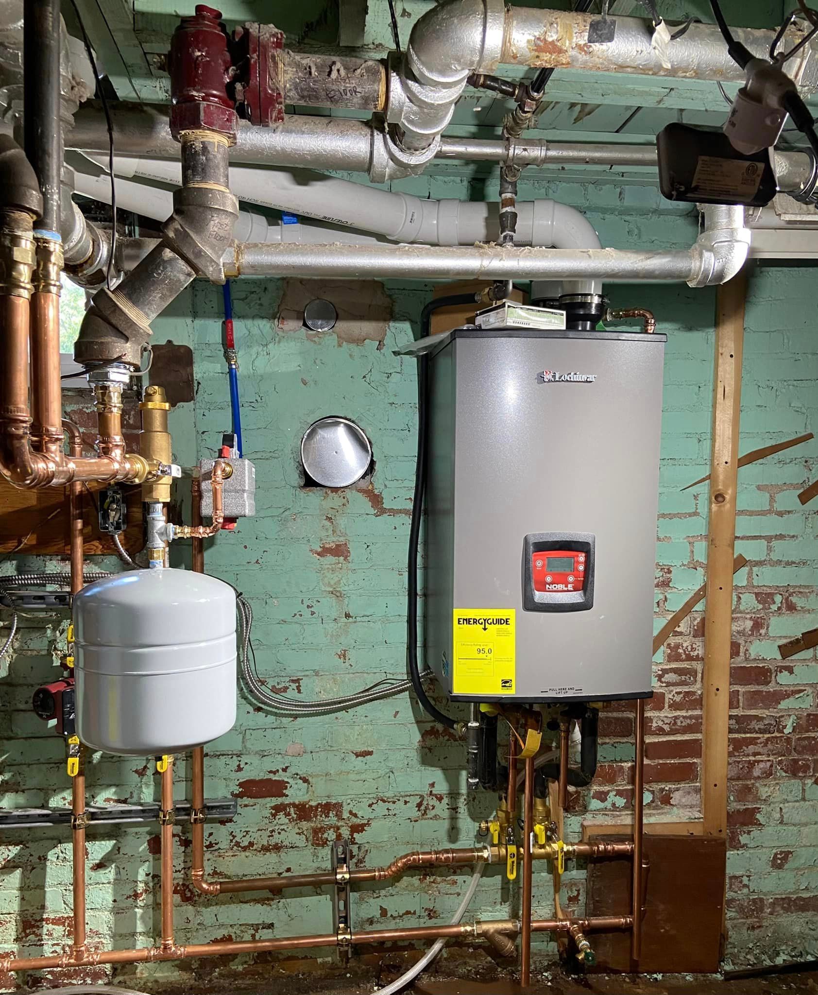 A water heater is sitting in a basement surrounded by pipes.