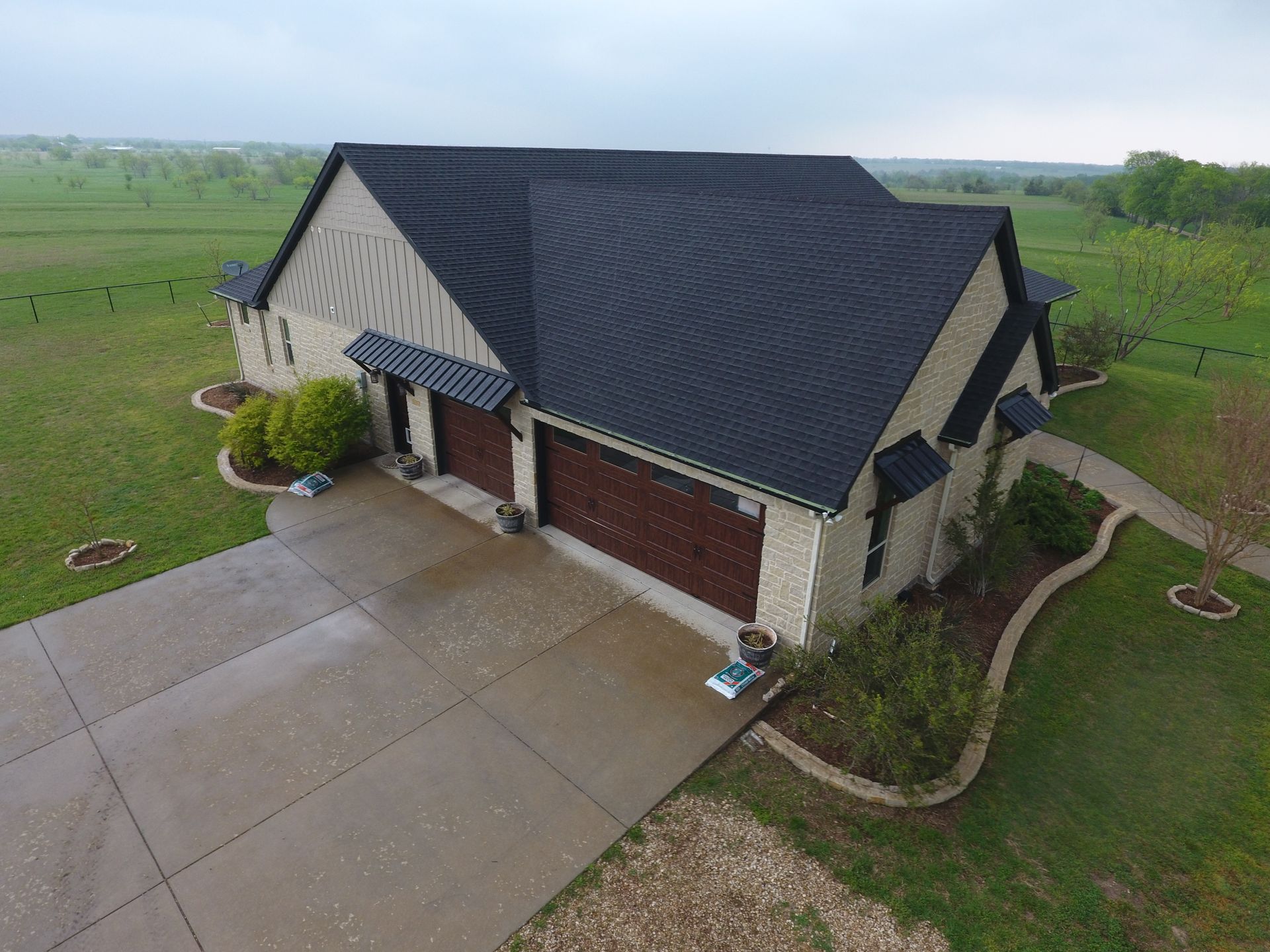 Aerial view of a suburban house with a dark roof, brick garage, and wide concrete driveway surrounded by green lawn