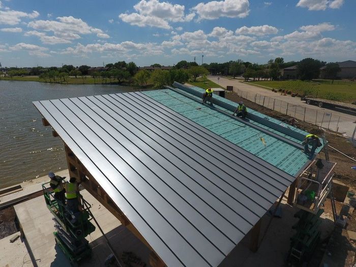Workers install silver metal roofing on a building frame by a river under a cloudy sky