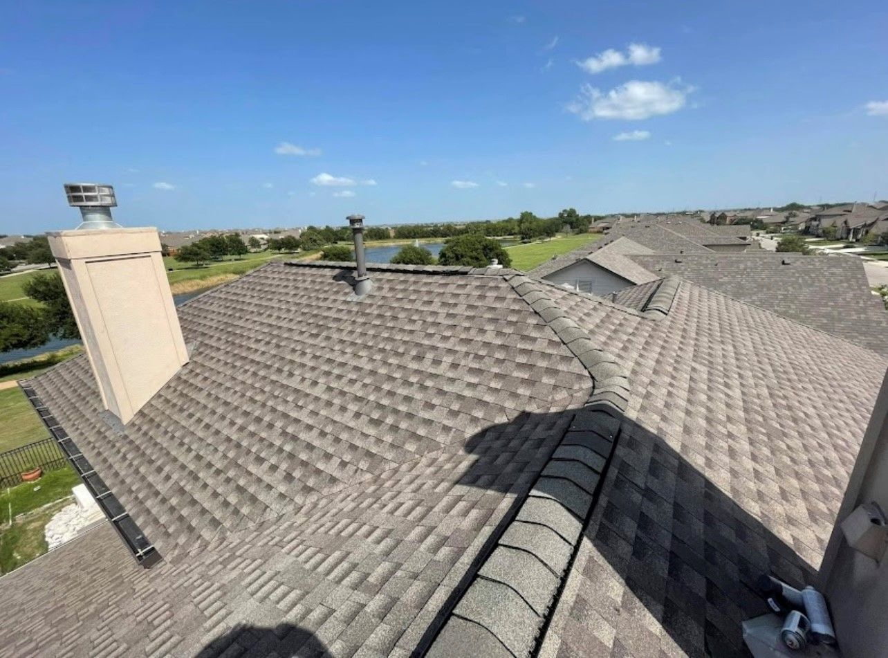 A rooftop with brown shingles and a beige chimney against a blue sky, overlooking a suburban neighborhood. A rooftop with brown shingles and a beige chimney against a blue sky, overlooking a suburban neighborhood.