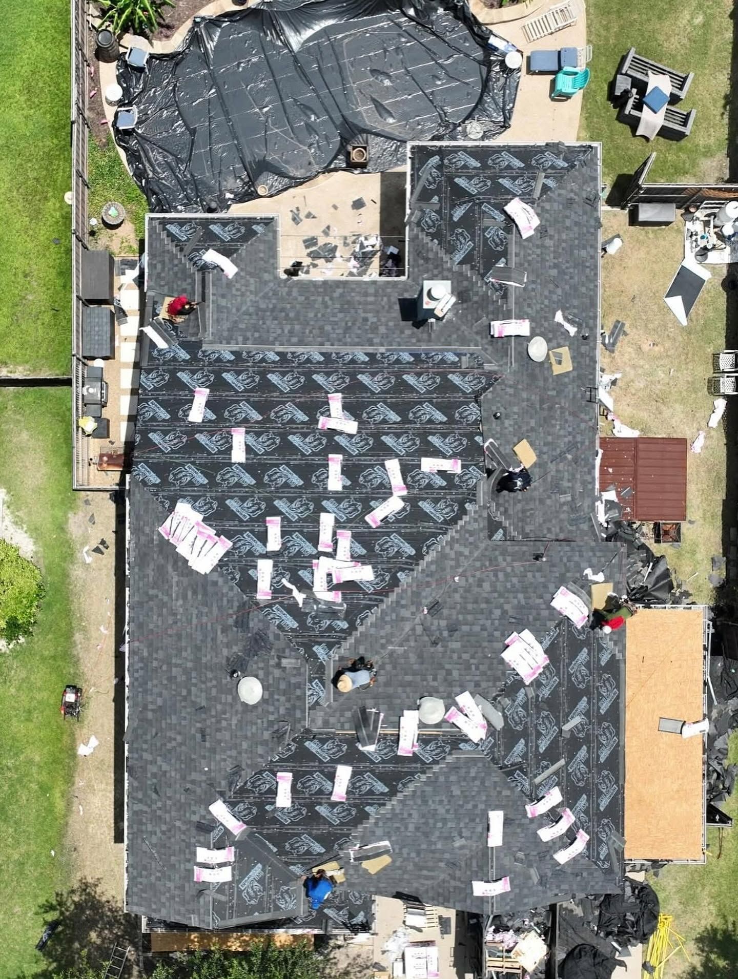 Aerial view of a roof under repair with black tarps, workers, and scattered equipment around the house.