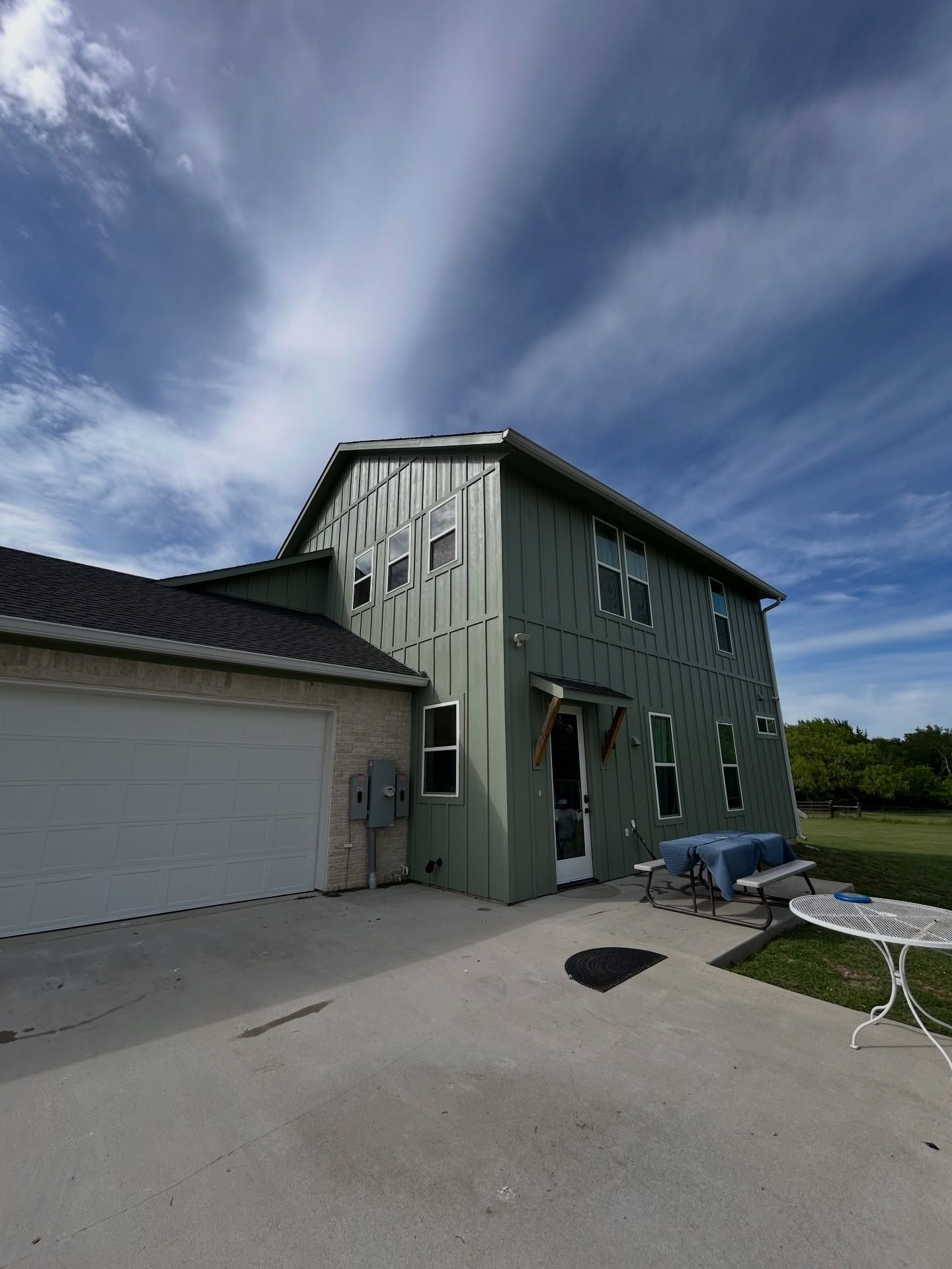 Green two-story house with white garage, concrete driveway, and lawn chairs under a blue sky
