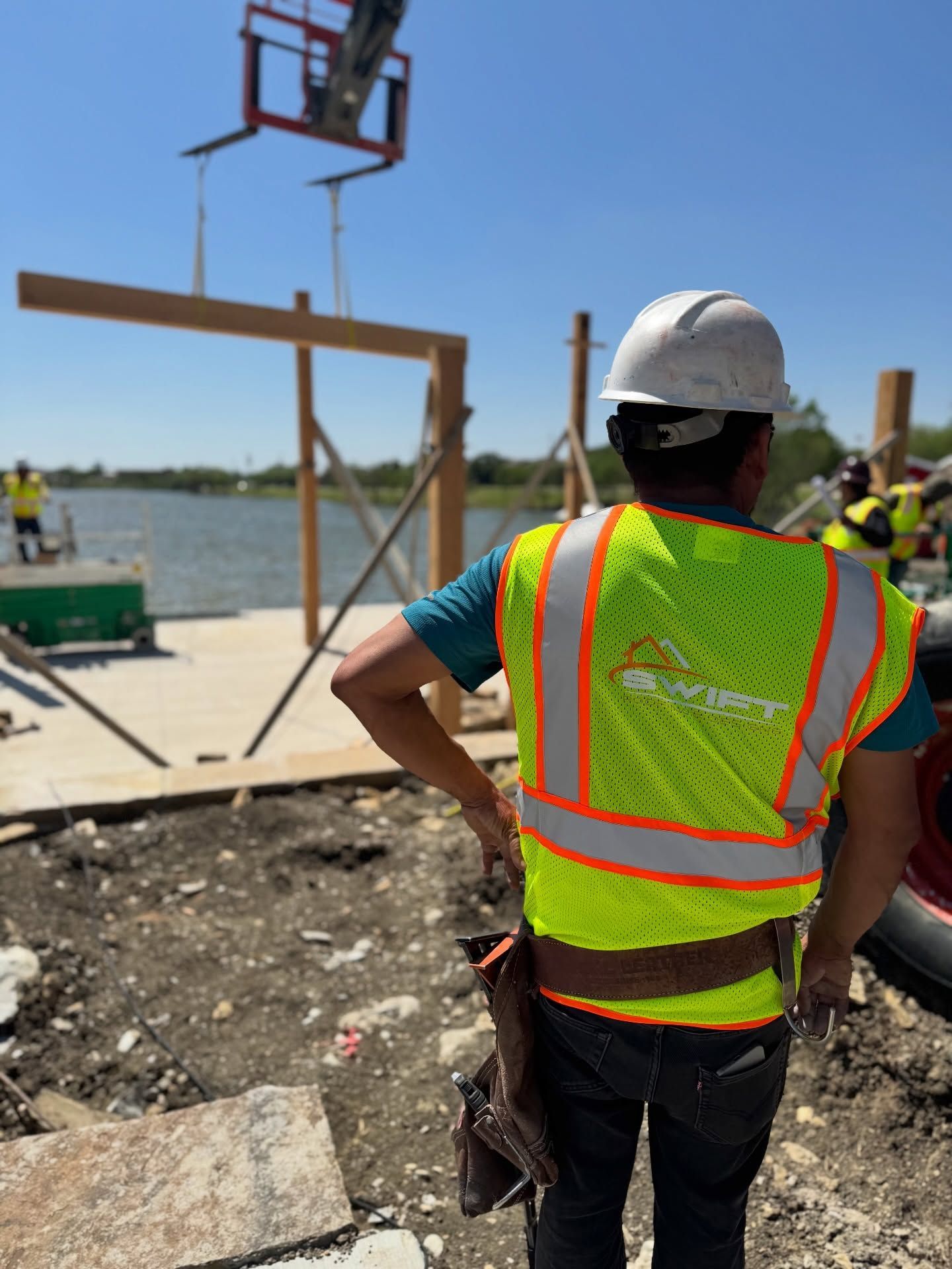 Construction worker in neon vest and hard hat overlooking a waterfront worksite with a crane lift