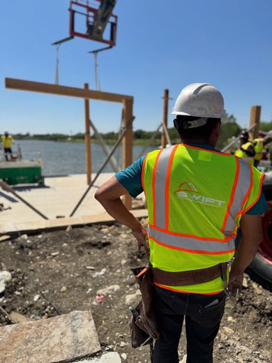 Construction worker in neon vest and hard hat overlooking a waterfront worksite with a crane lift