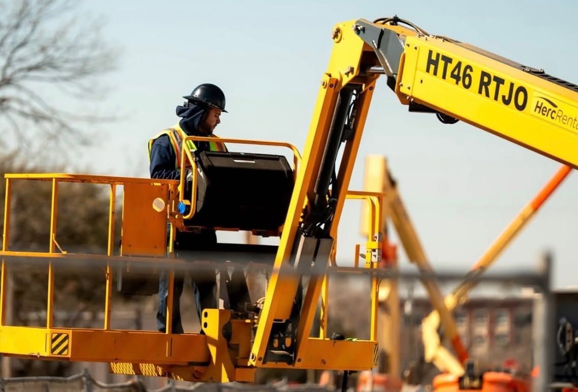 Worker in a yellow lift basket operating a yellow boom arm on a construction site