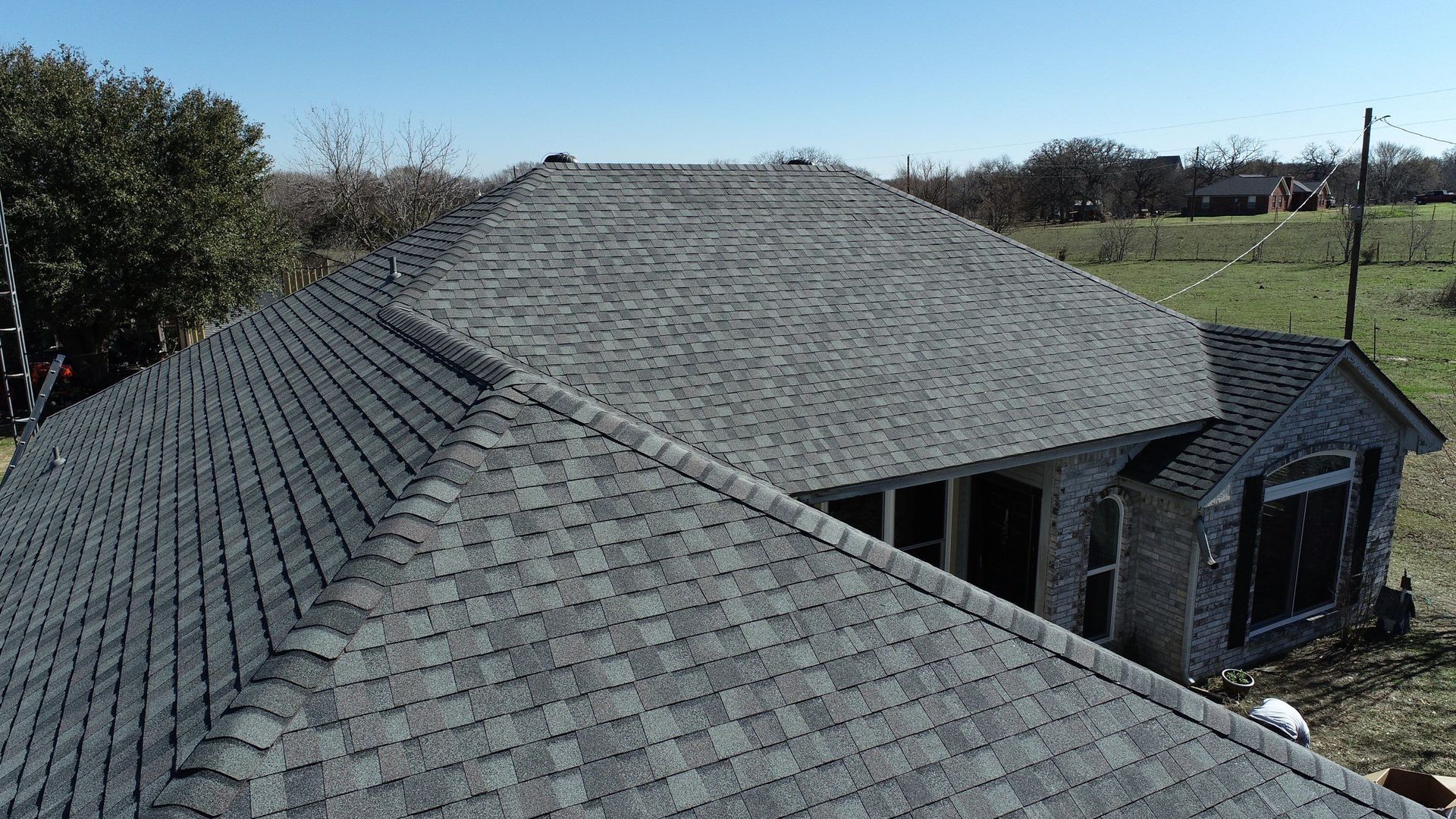 Aerial view of a gray-shingled roof on a house in a rural area under a clear sky.