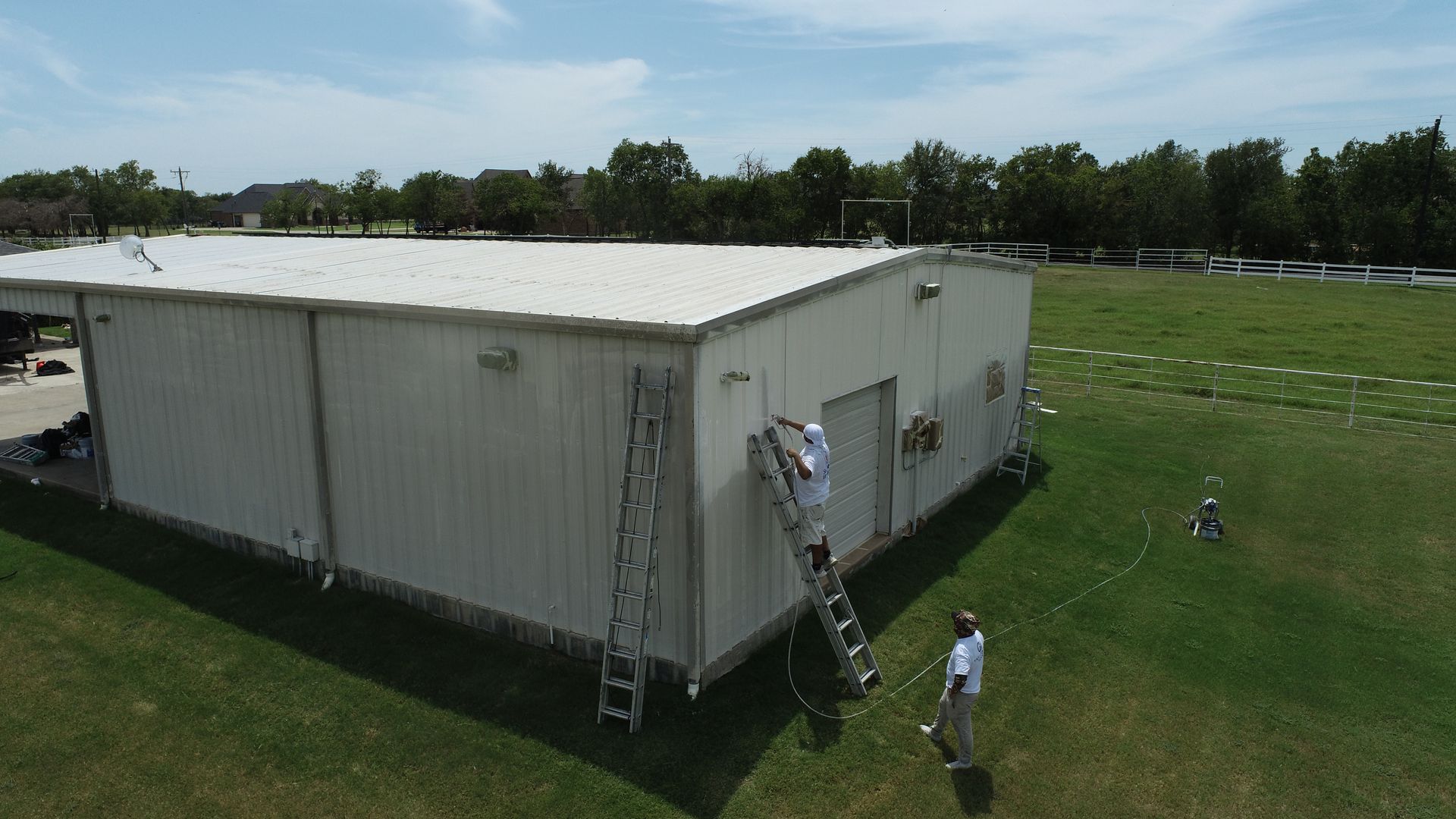 Two people paint the side of a white metal building with ladders on a grassy field on a sunny day. Two people paint the side of a white metal building with ladders on a grassy field on a sunny day.