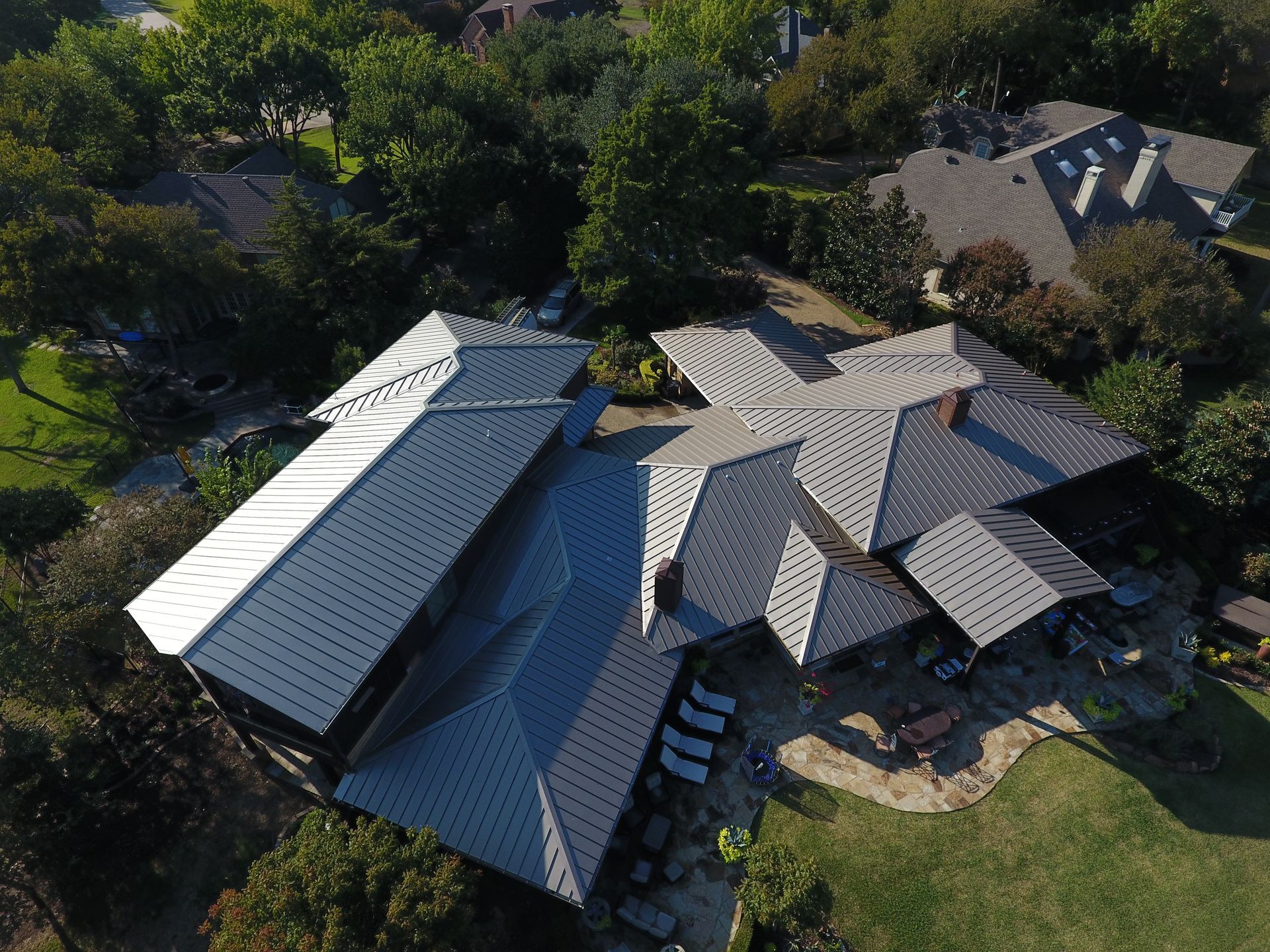 Aerial view of a house with multiple gray metal roofs surrounded by green trees and lawn.