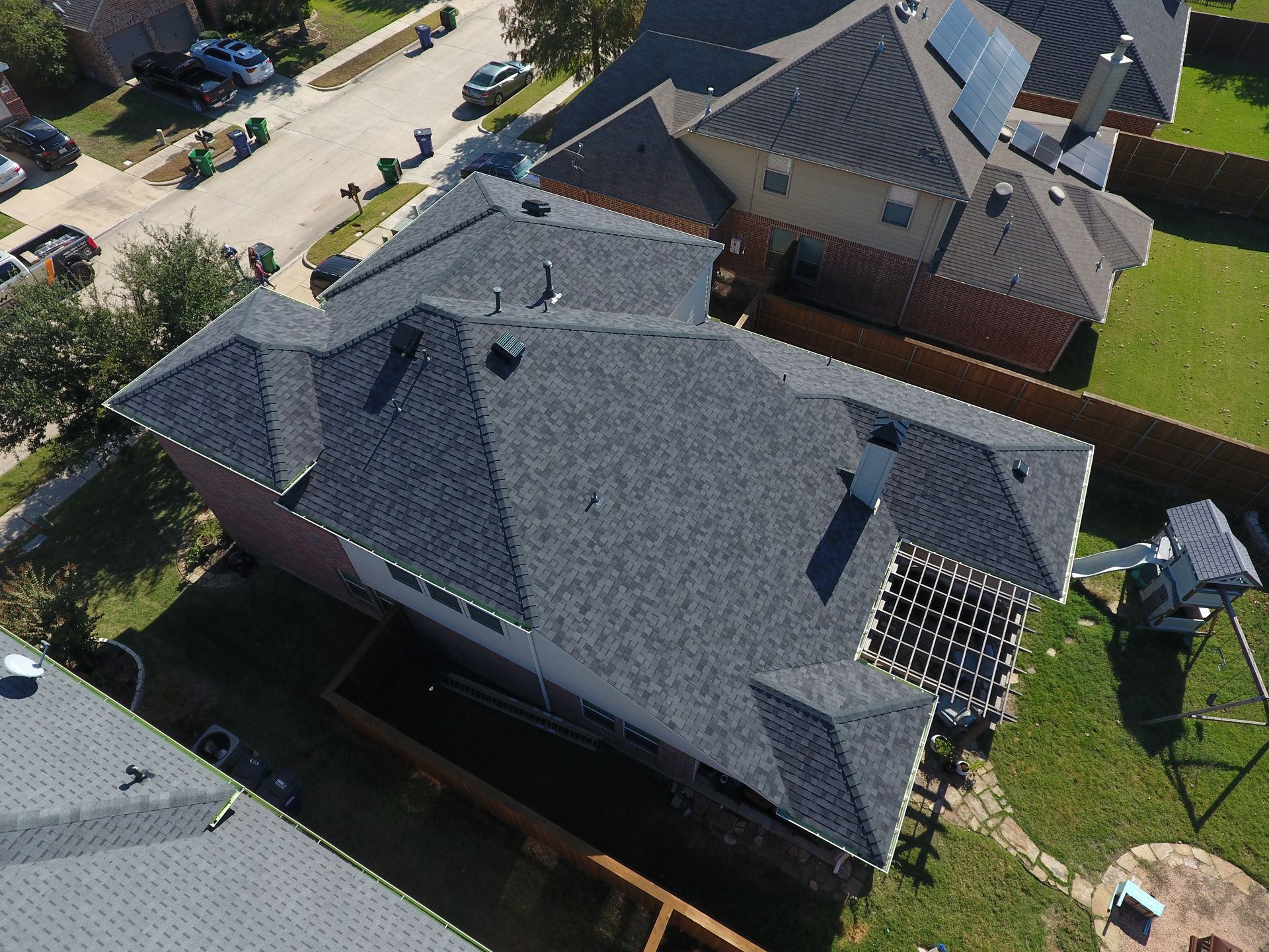 Overhead view of a house with a dark gray roof surrounded by green lawn and neighboring houses.