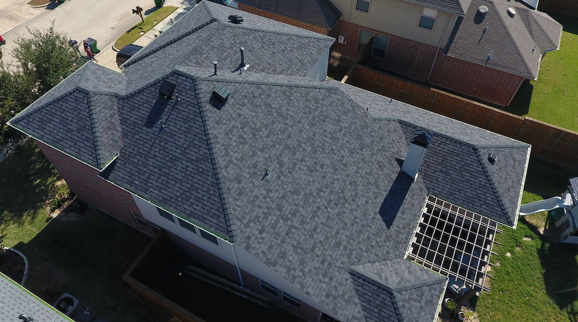 Overhead view of a dark gray shingle roof on a two-story house with chimneys and a small pergola.