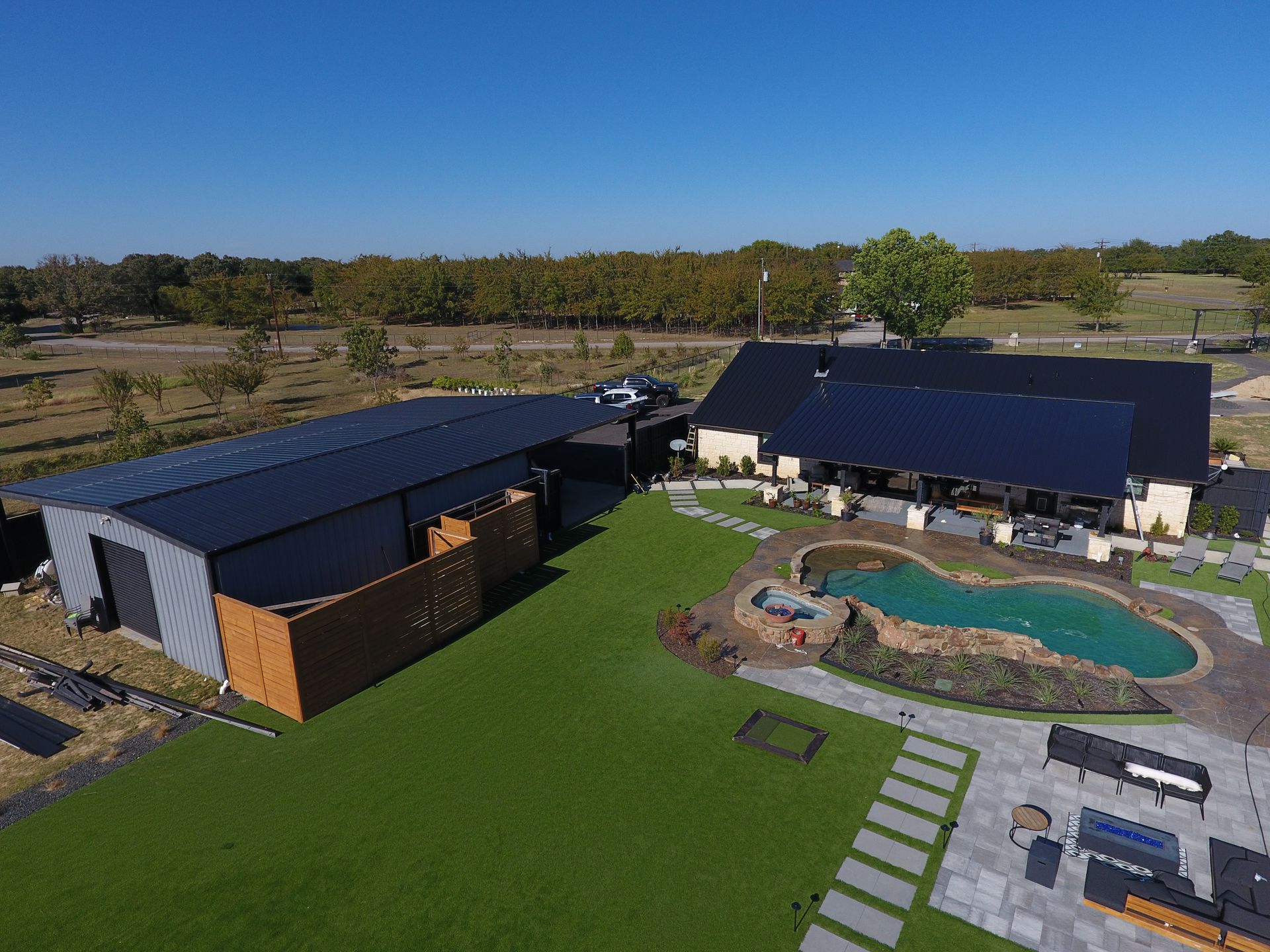 Aerial view of a modern home with pool, landscaping, and a large black roofed structure on a green lawn.