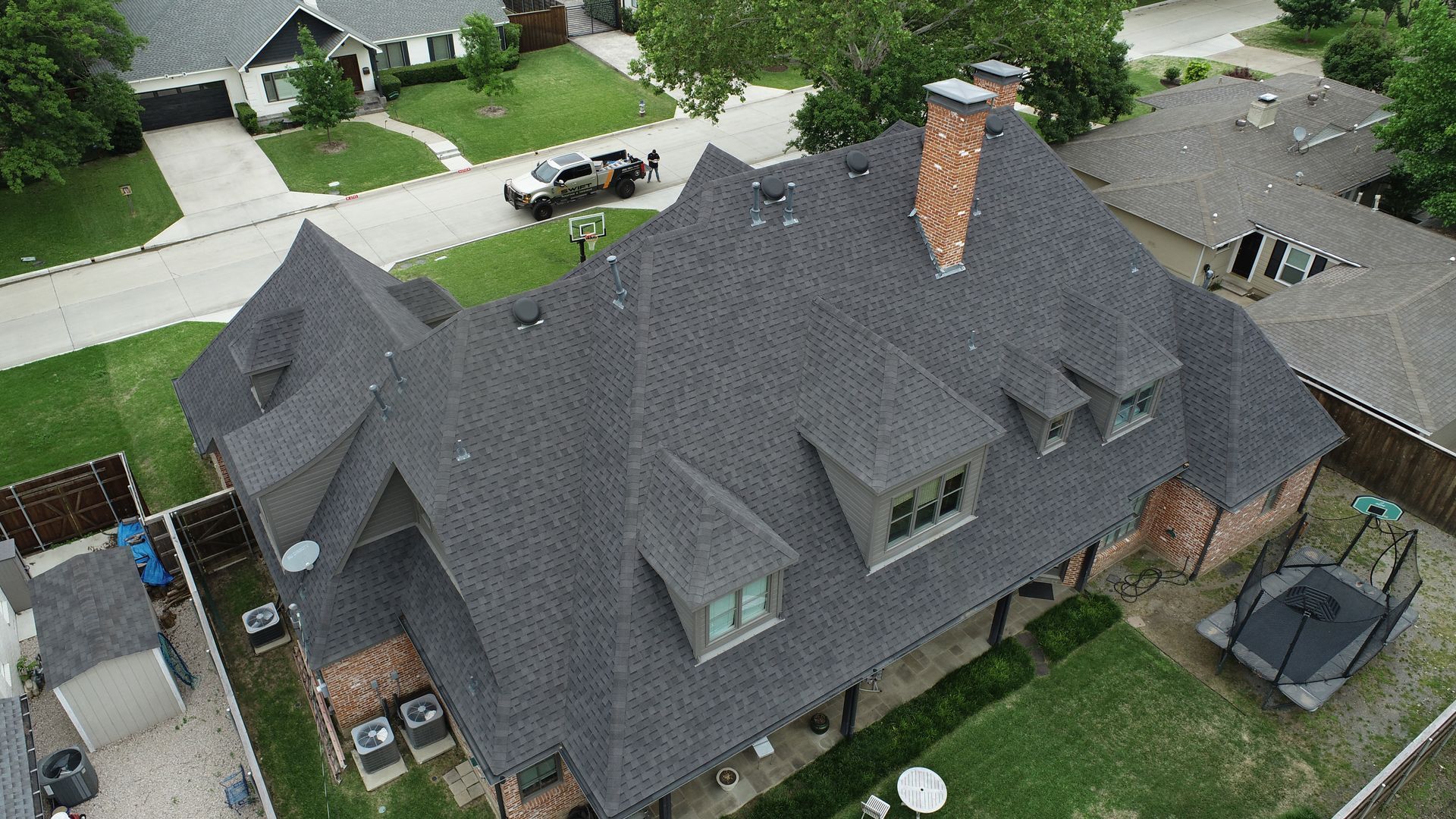Aerial view of a dark gray roof with a brick chimney and multiple dormer windows on a house surrounded by green grass.