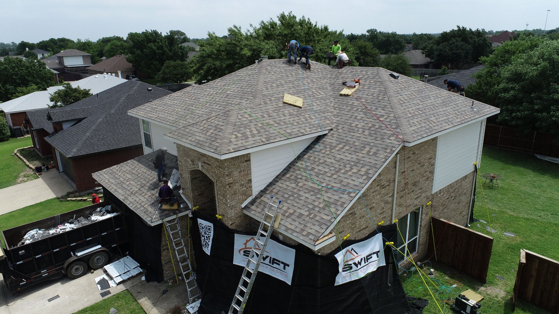 Roofers working on a two-story home, removing old shingles and installing new ones; a dumpster is nearby.