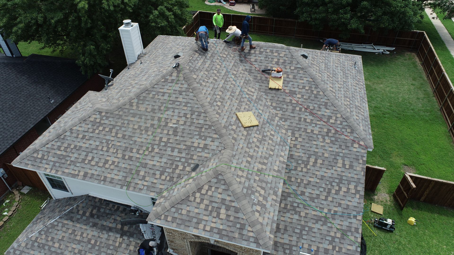 Workers on a roof replacing shingles. Green lawn surrounds the house, with a white chimney.