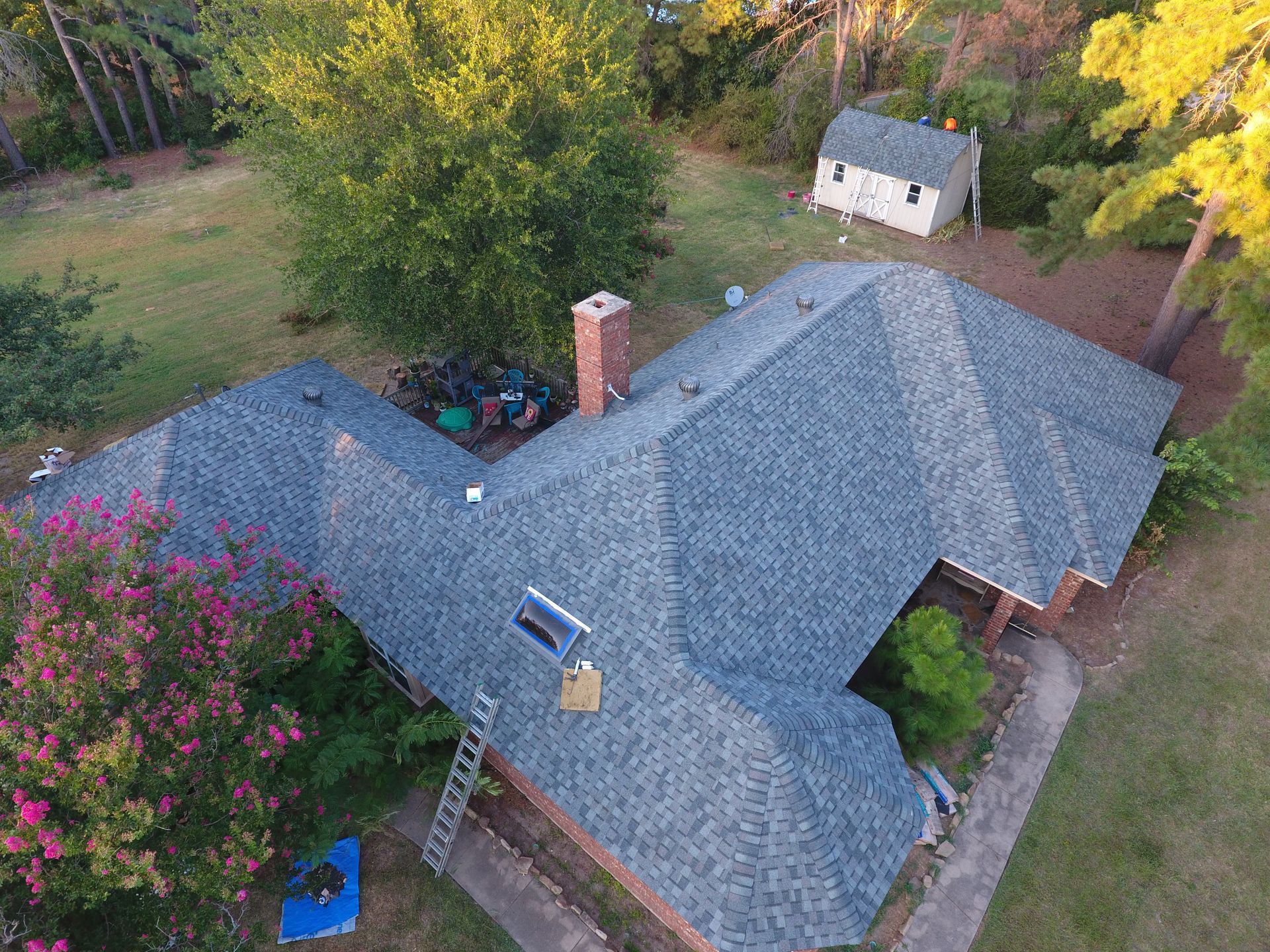Aerial view of a house with a gray roof, chimney, and a small shed. Green trees surround the home.