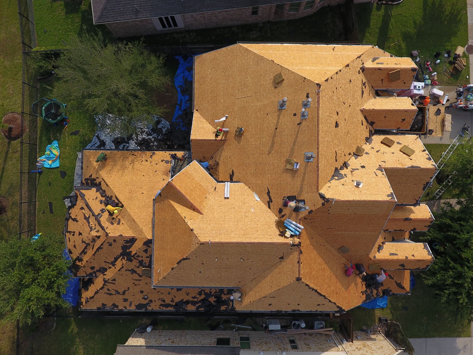 Aerial view of a house roof partially covered with brown shingles, and blue tarps, suggesting storm damage.