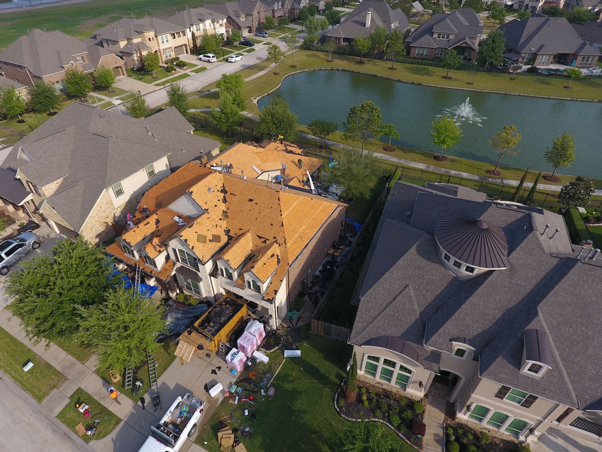 Aerial view of a house with significant roof damage, debris in the yard, near a pond and other houses.