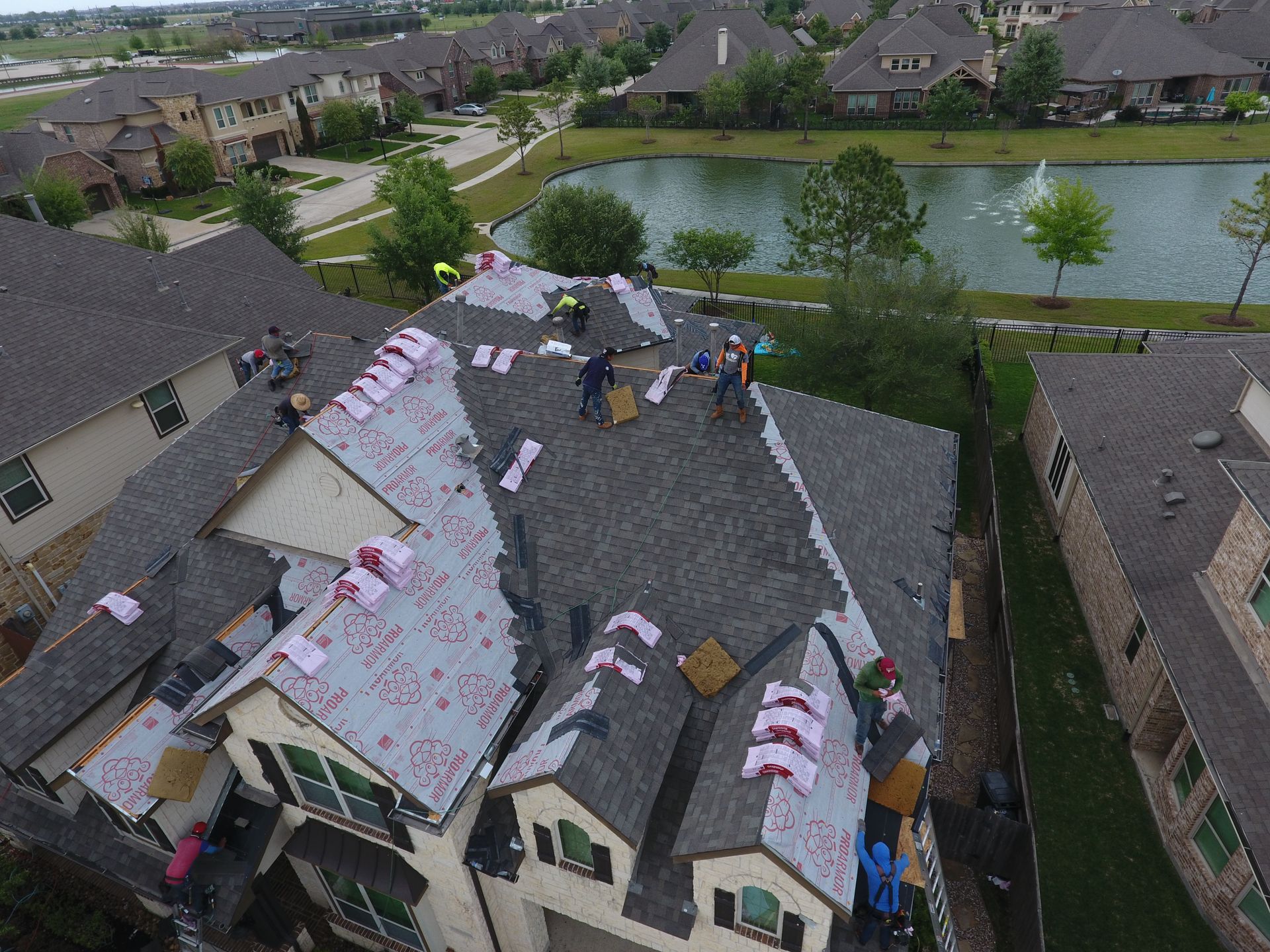 Aerial view of roof damage with workers repairing shingles in a suburban neighborhood near a pond
