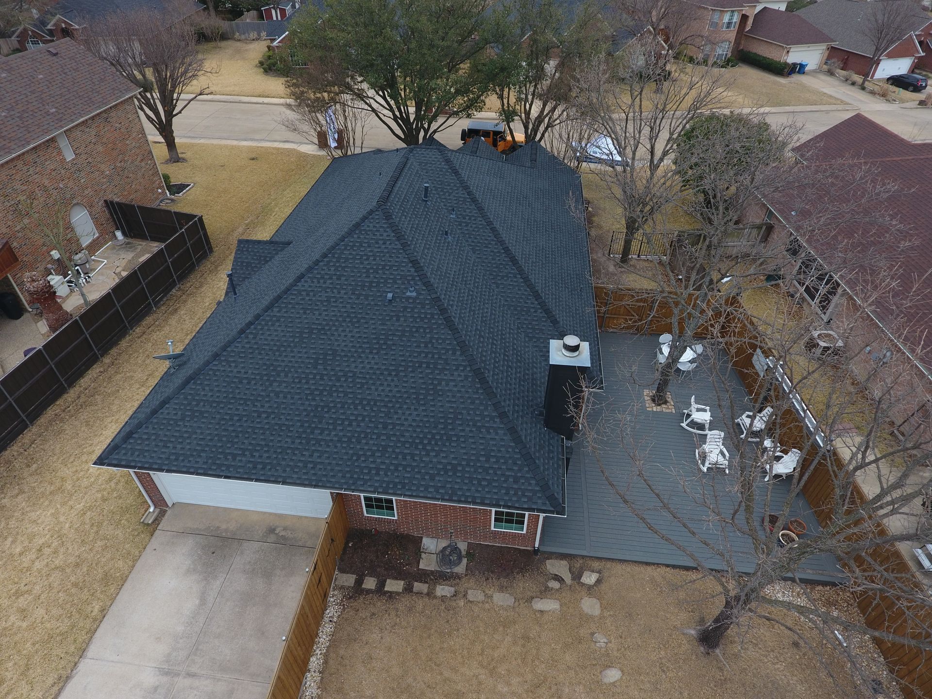 Aerial view of a house with a dark gray shingle roof, patio, and driveway.