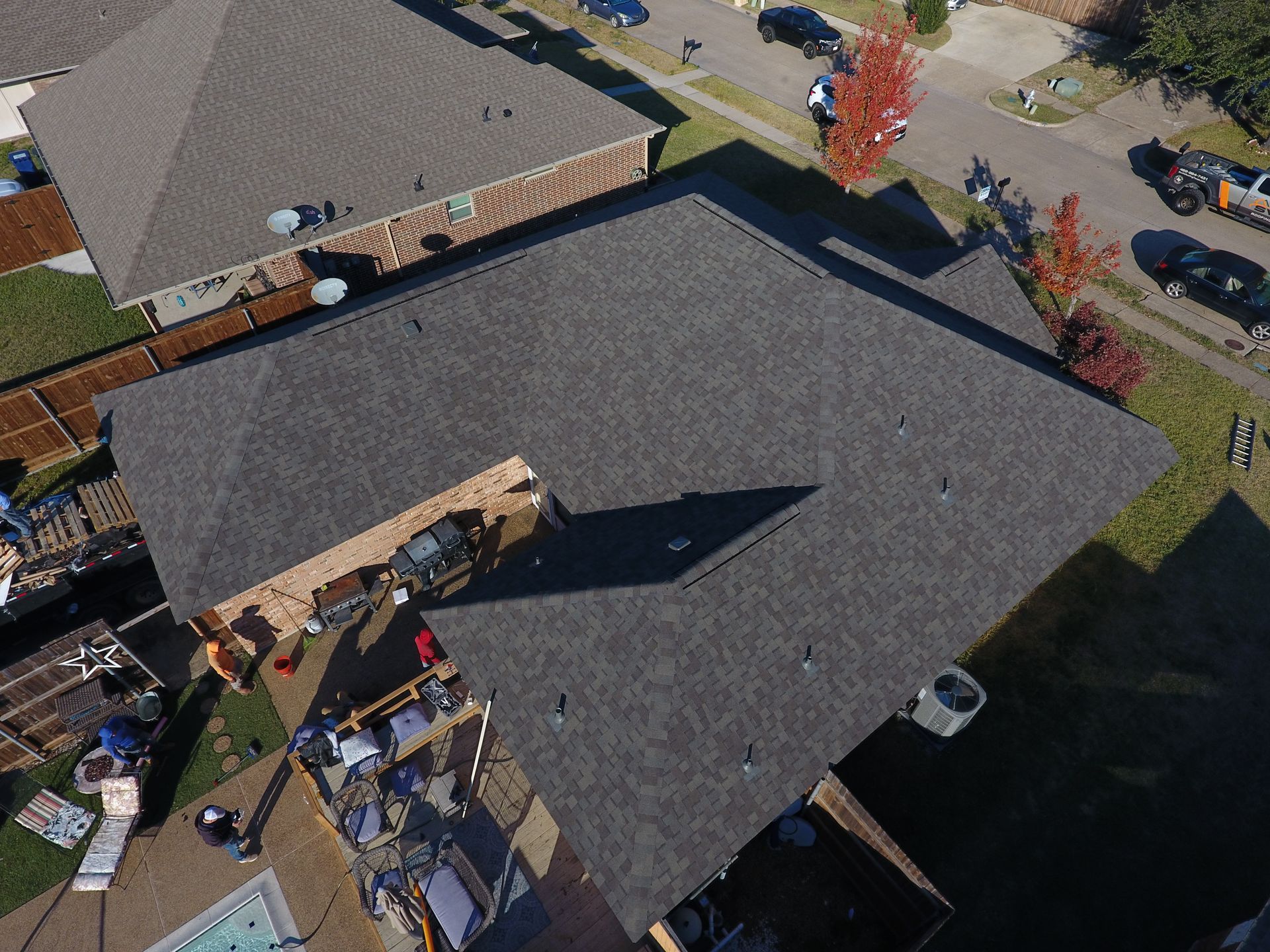Aerial view of a dark-roofed house with a driveway, patio furniture, and a parked car beside a road
