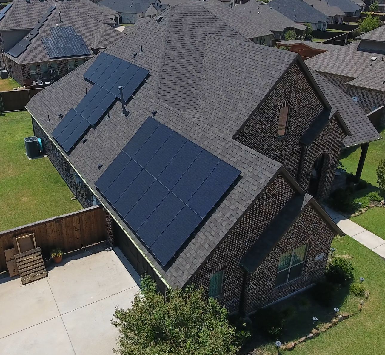 Aerial view of a suburban house with dark roof shingles and solar panels in the driveway-side roof area.