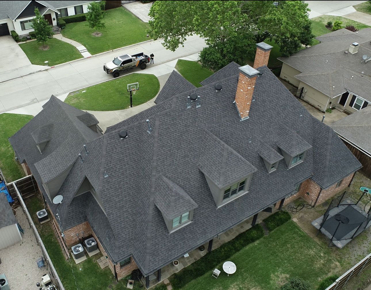 Aerial view of a dark shingled roof on a large brick house. A car drives on the street in front of the house.