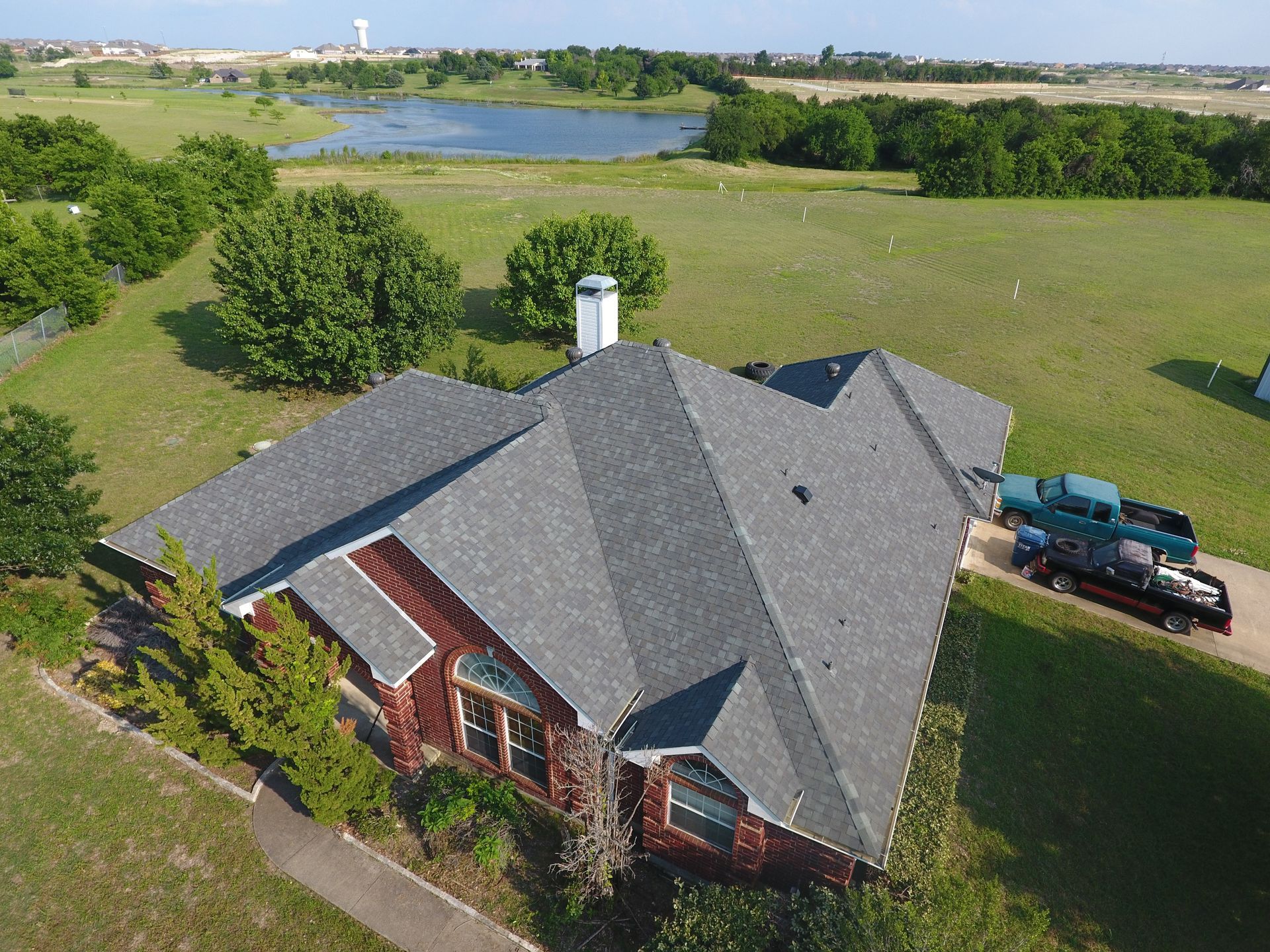 Drone shot of a red-sided house with a gray roof, green lawn, pond, and blue pickup trucks.