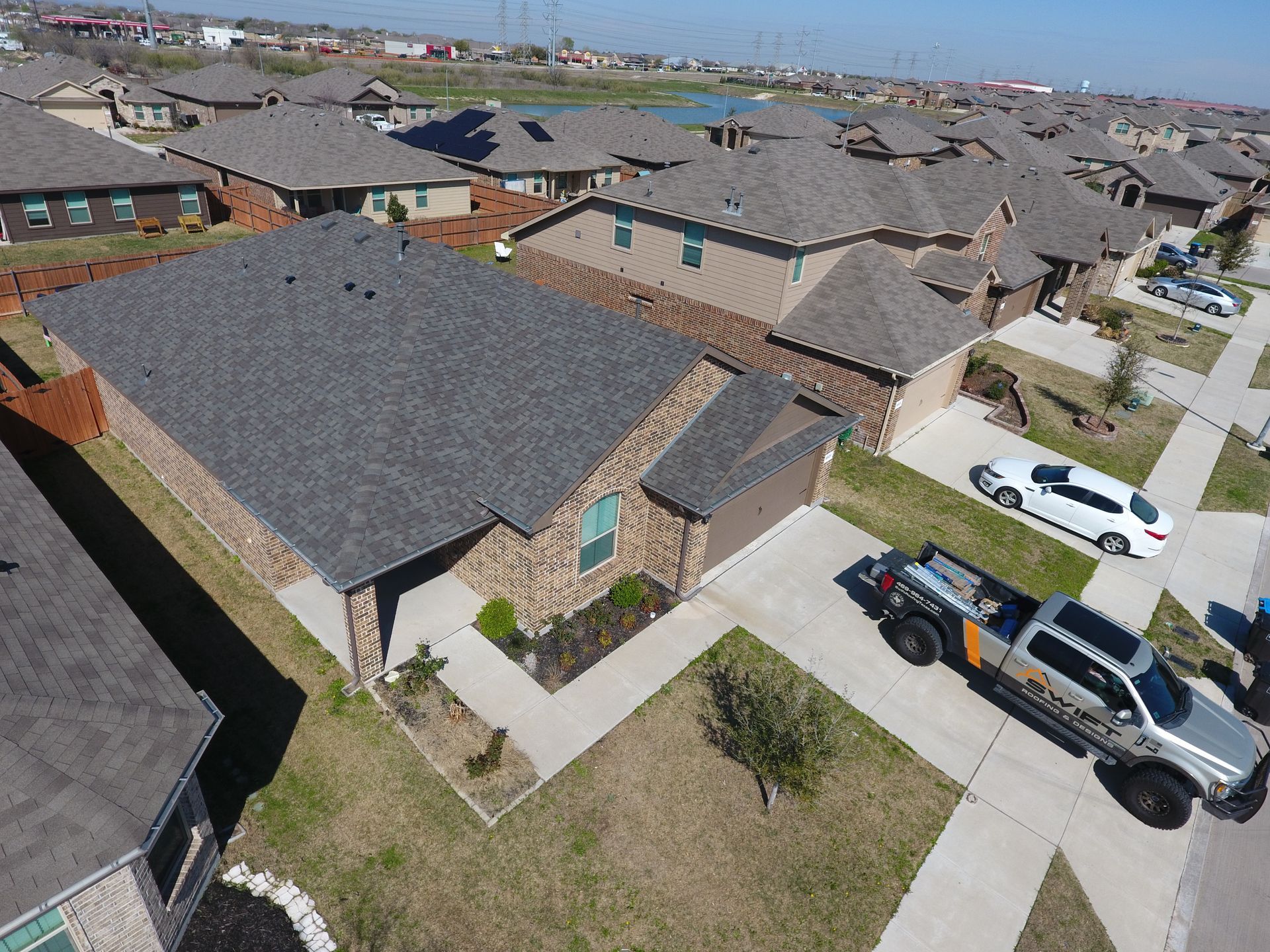 Aerial view of suburban houses with dark gray roofs and parked vehicles on the street.