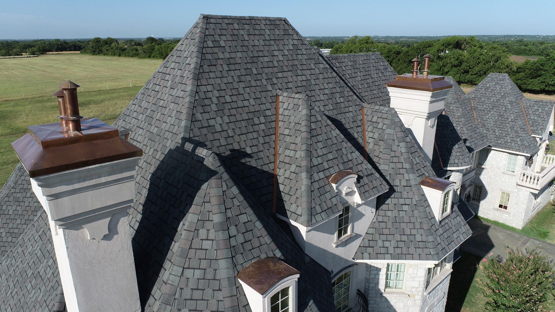 Gray shingled roof with copper accents on a light-colored building, surrounded by green fields.