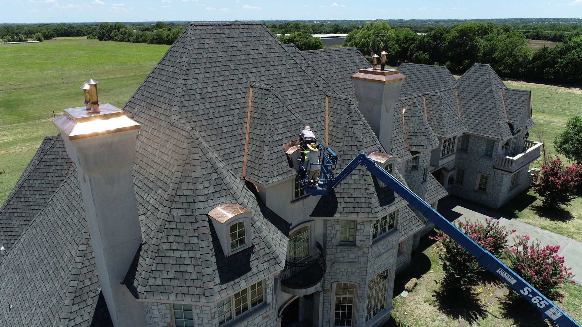 Workers on a lift repairing a gray tiled roof of a large house with copper accents; sunny day.