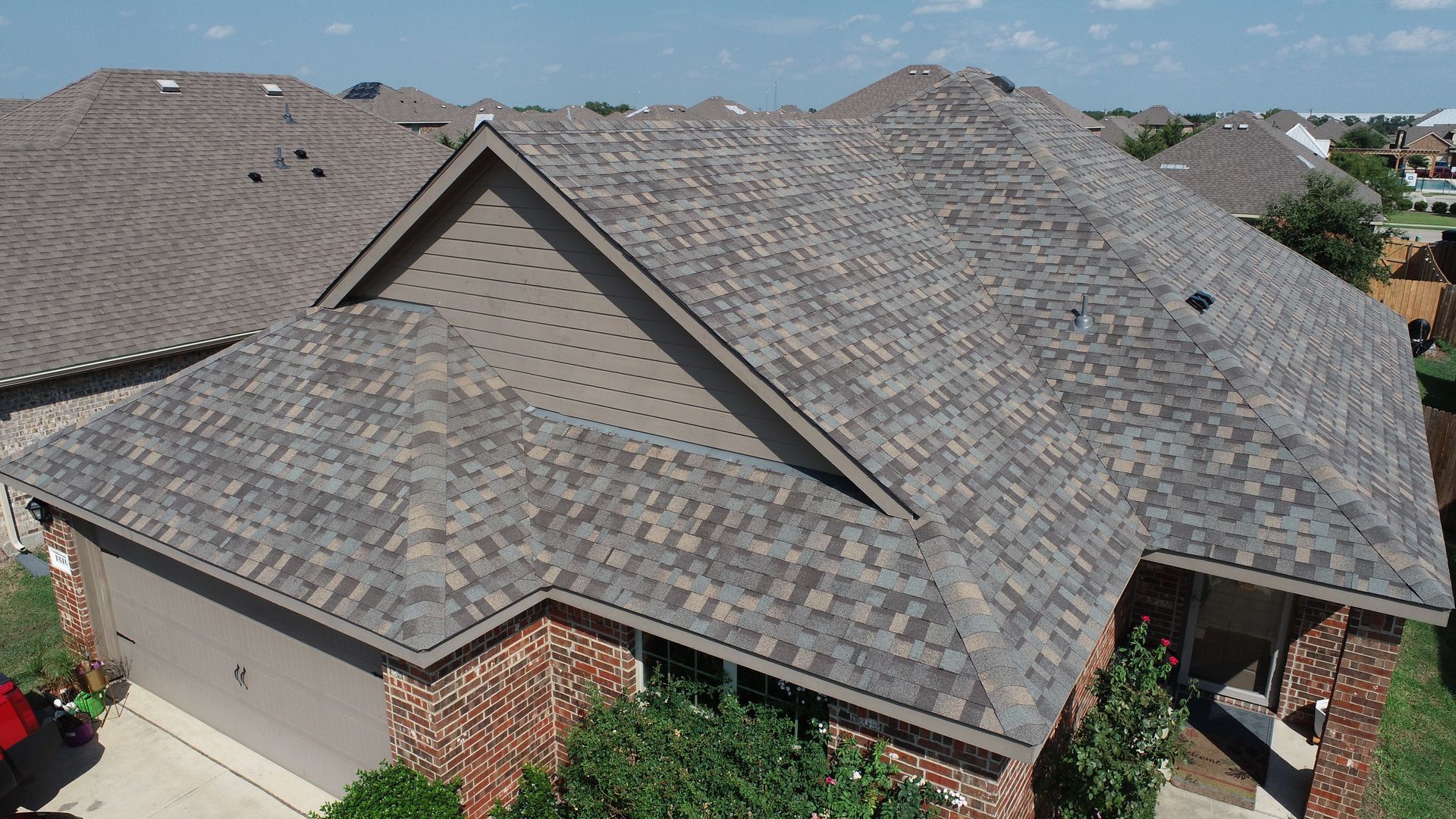 Aerial view of a brick house with a gray shingle roof and surrounding greenery.