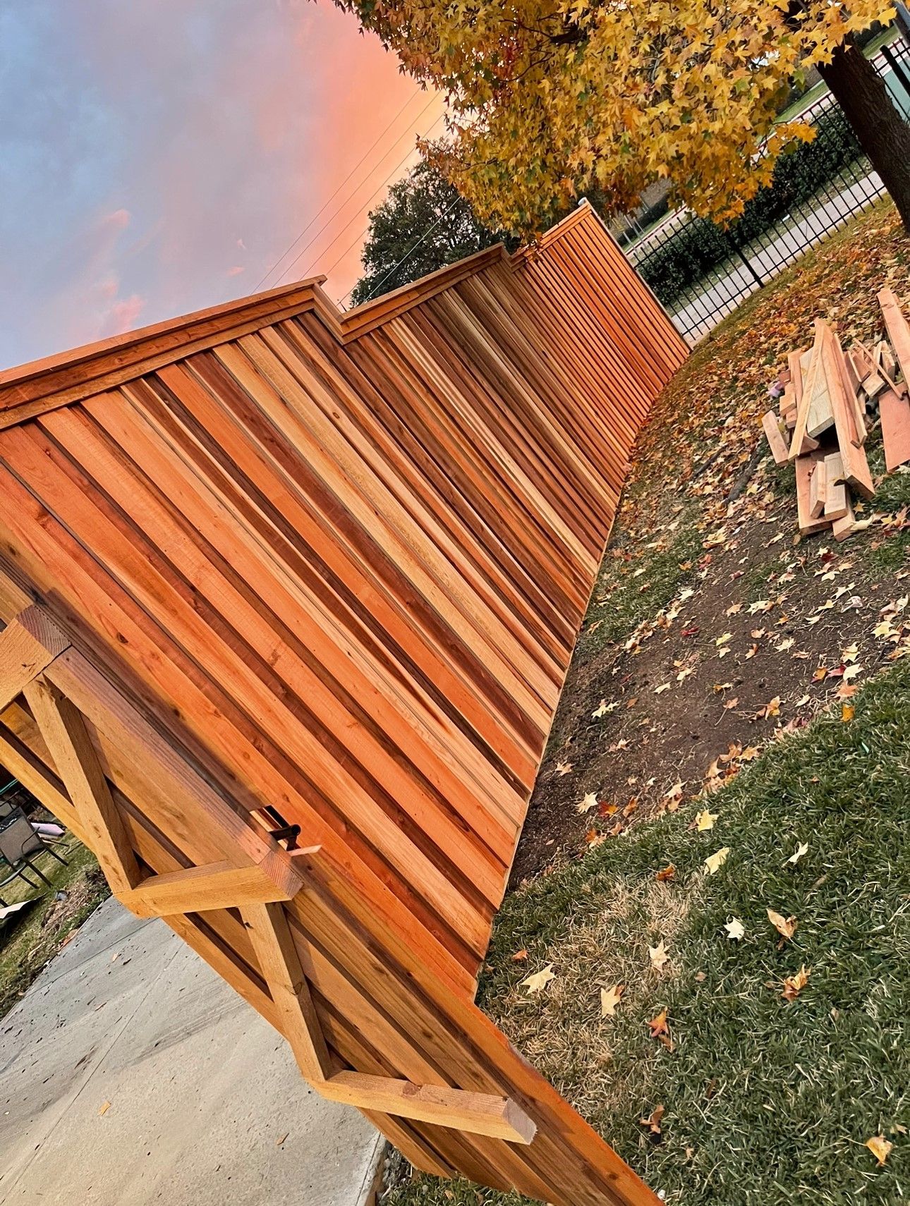 Wooden fence with a warm stain, grass, fallen leaves, and a colorful sunset sky.