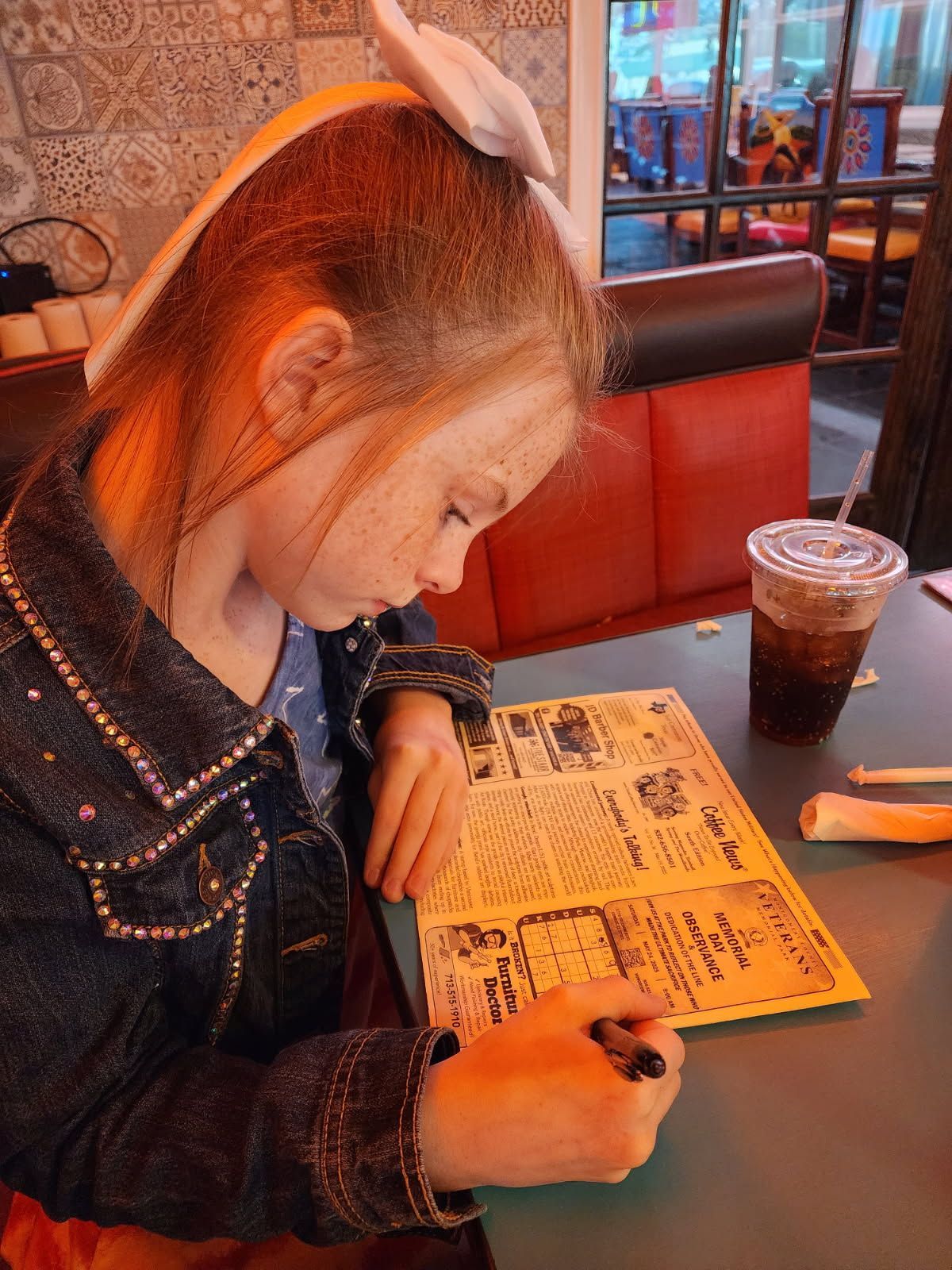 Girl with bow in hair, writing on a menu in a restaurant, drink and red booth in background.