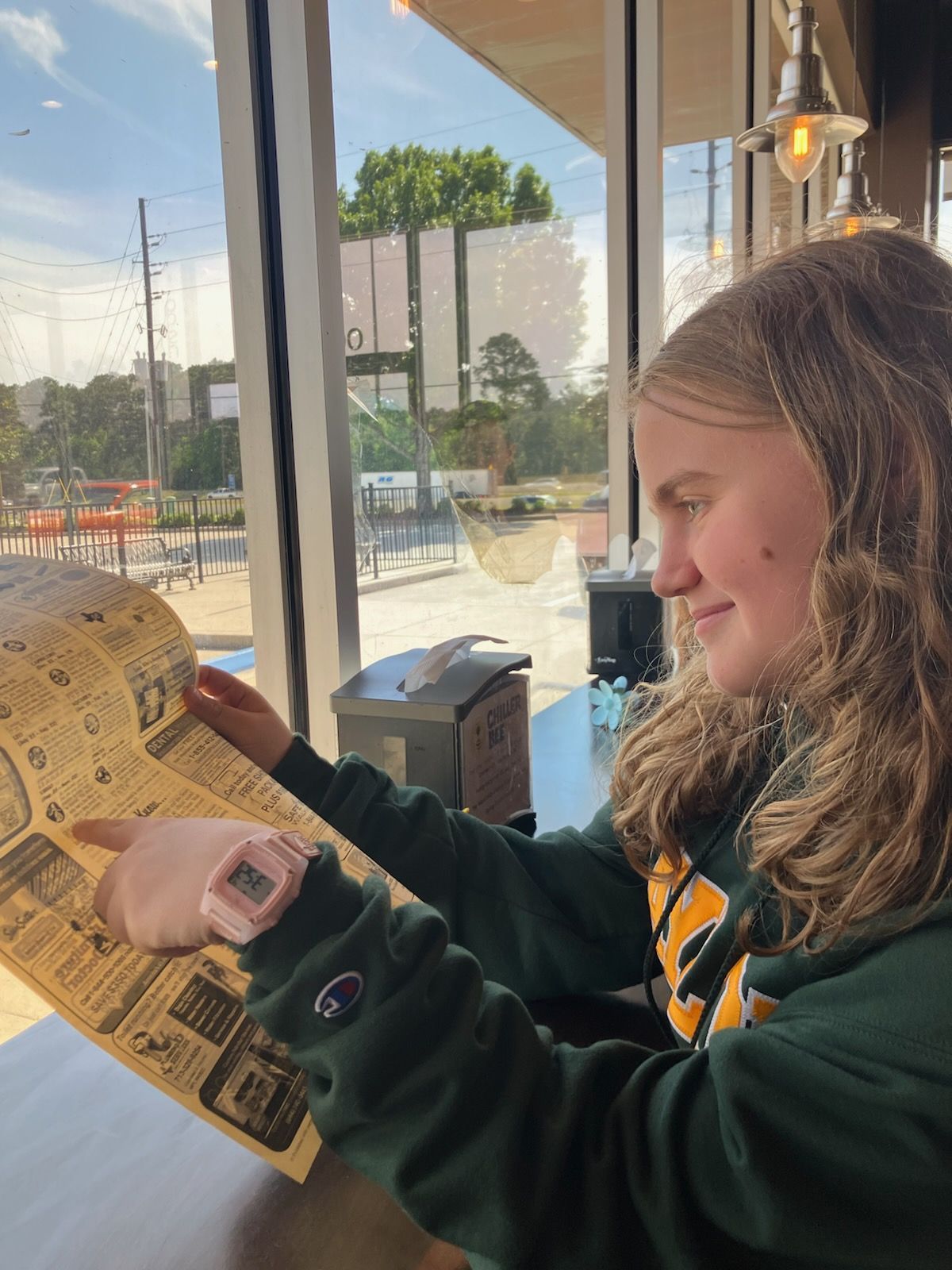 Person in a green sweatshirt points to a menu at a table by a window; sunny outdoor setting.