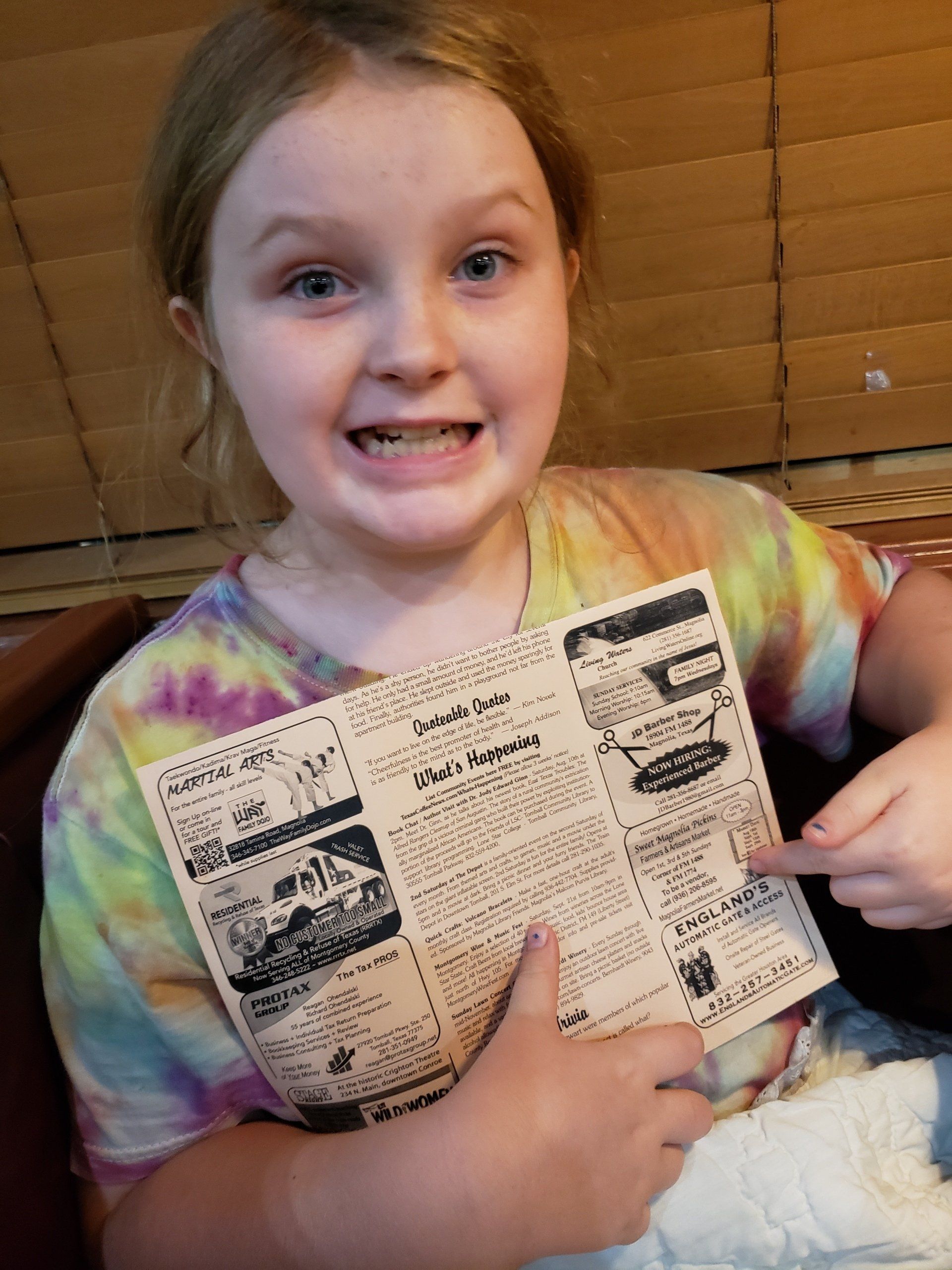 A young girl in a tie dye shirt is holding a newspaper