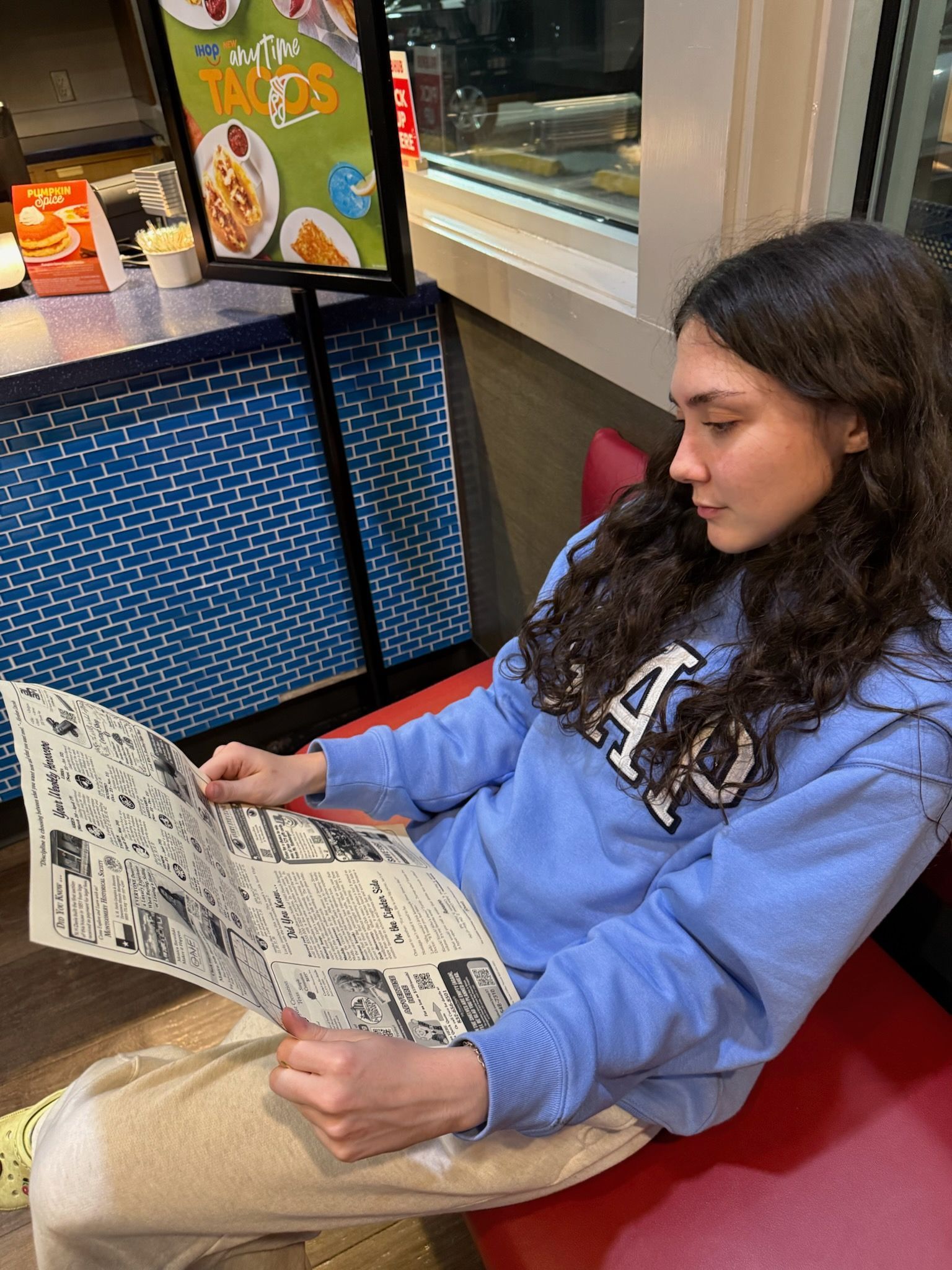 Woman in blue sweatshirt seated, reading a newspaper in a restaurant booth, with a taco sign visible.