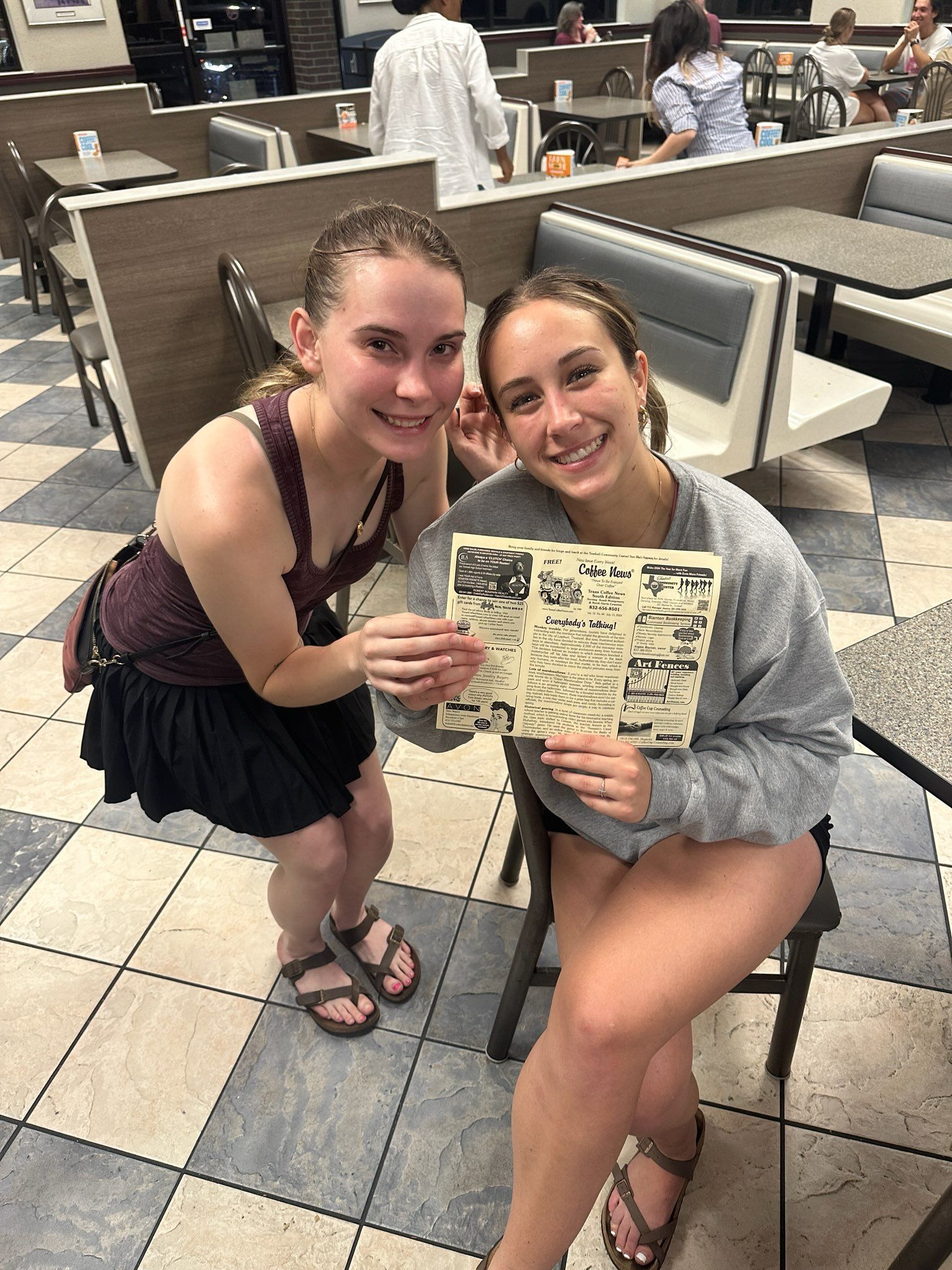 Two young women, one seated, hold a menu in a diner. They smile at the camera. Black and white tiled floor.