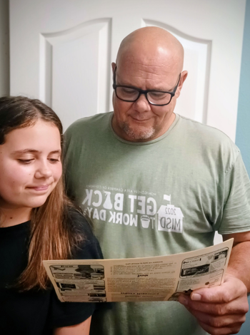 A man and a girl looking at a vintage advertisement, both smiling. The man wears glasses and a green shirt.