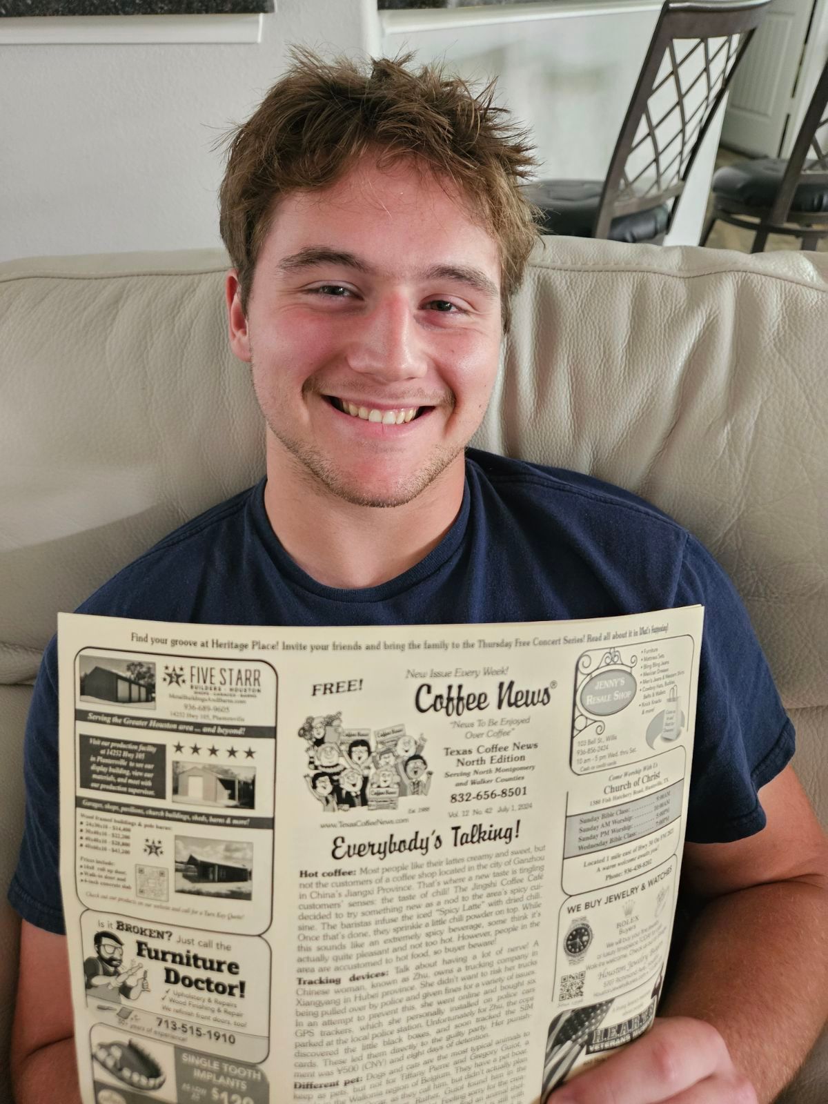 Man smiling, holding a vintage newspaper, seated on a beige couch indoors.