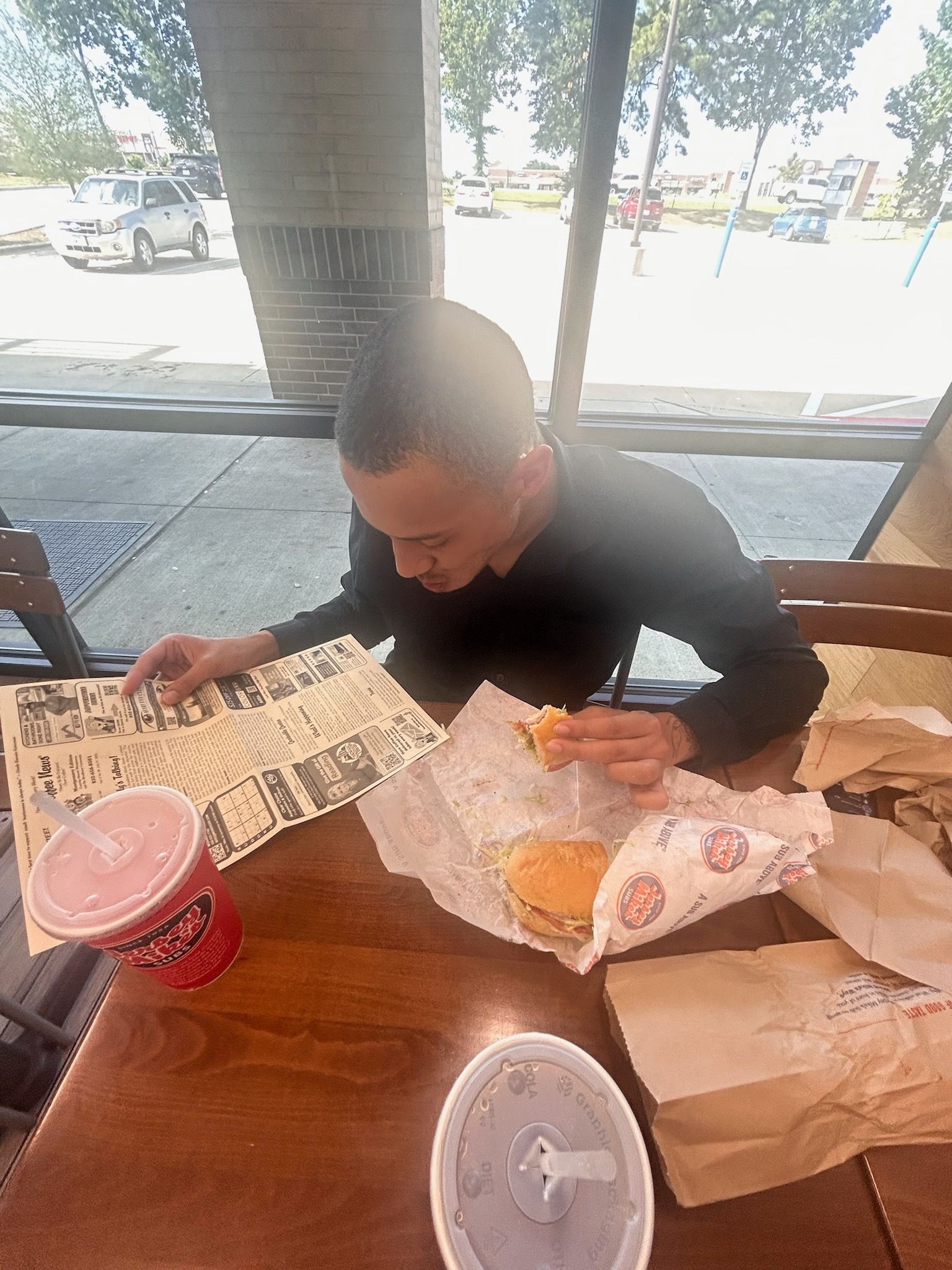 Person seated at table, eating burger and reading. Fast food setting with drink.