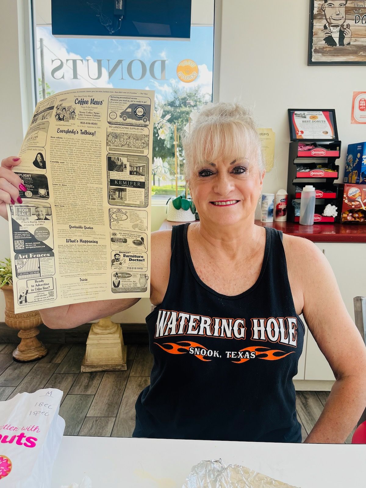 Woman in black tank top holds old newspaper in donut shop.