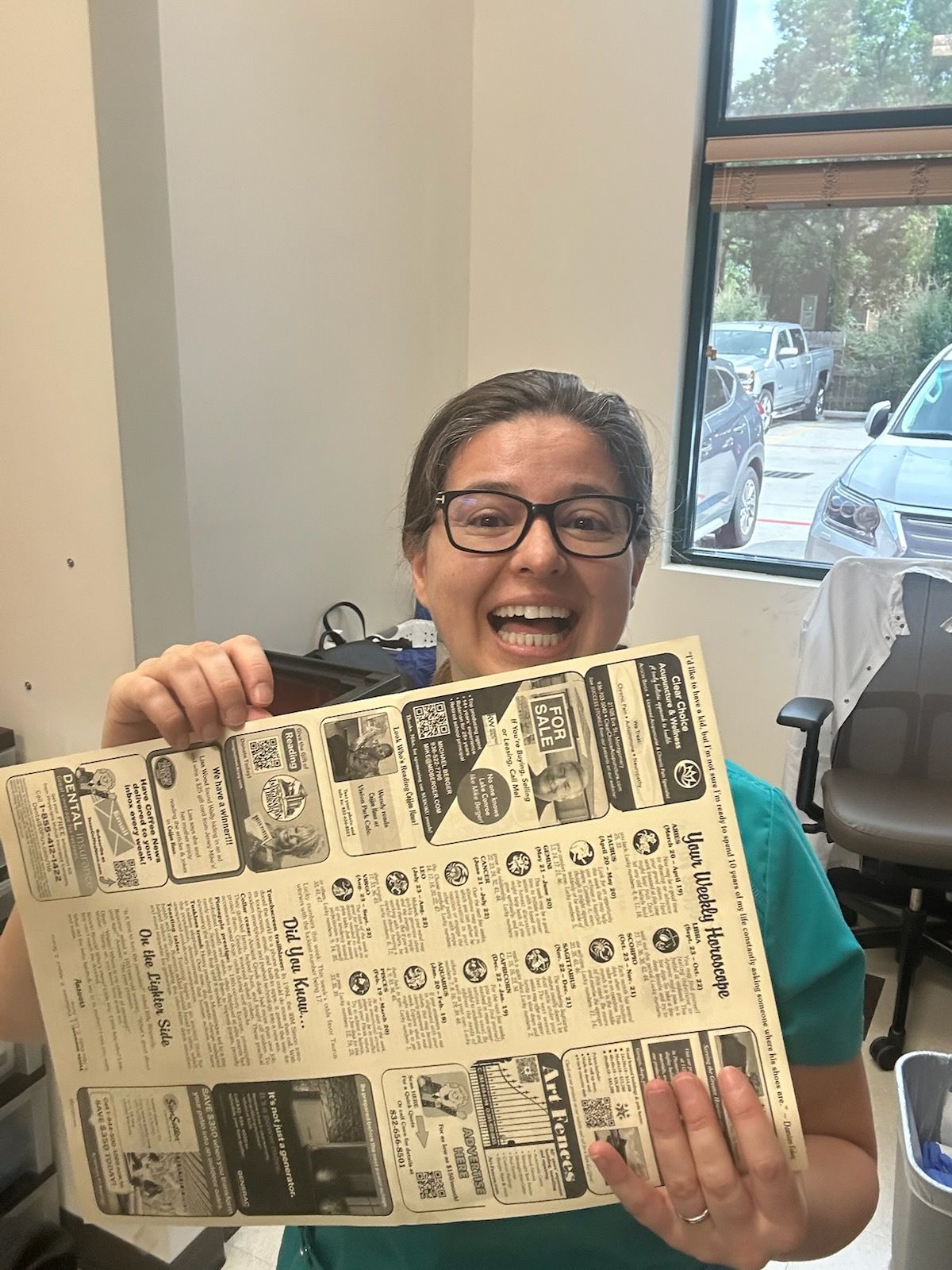 Woman holding a newspaper ad, smiling widely. Wearing glasses and a teal shirt.