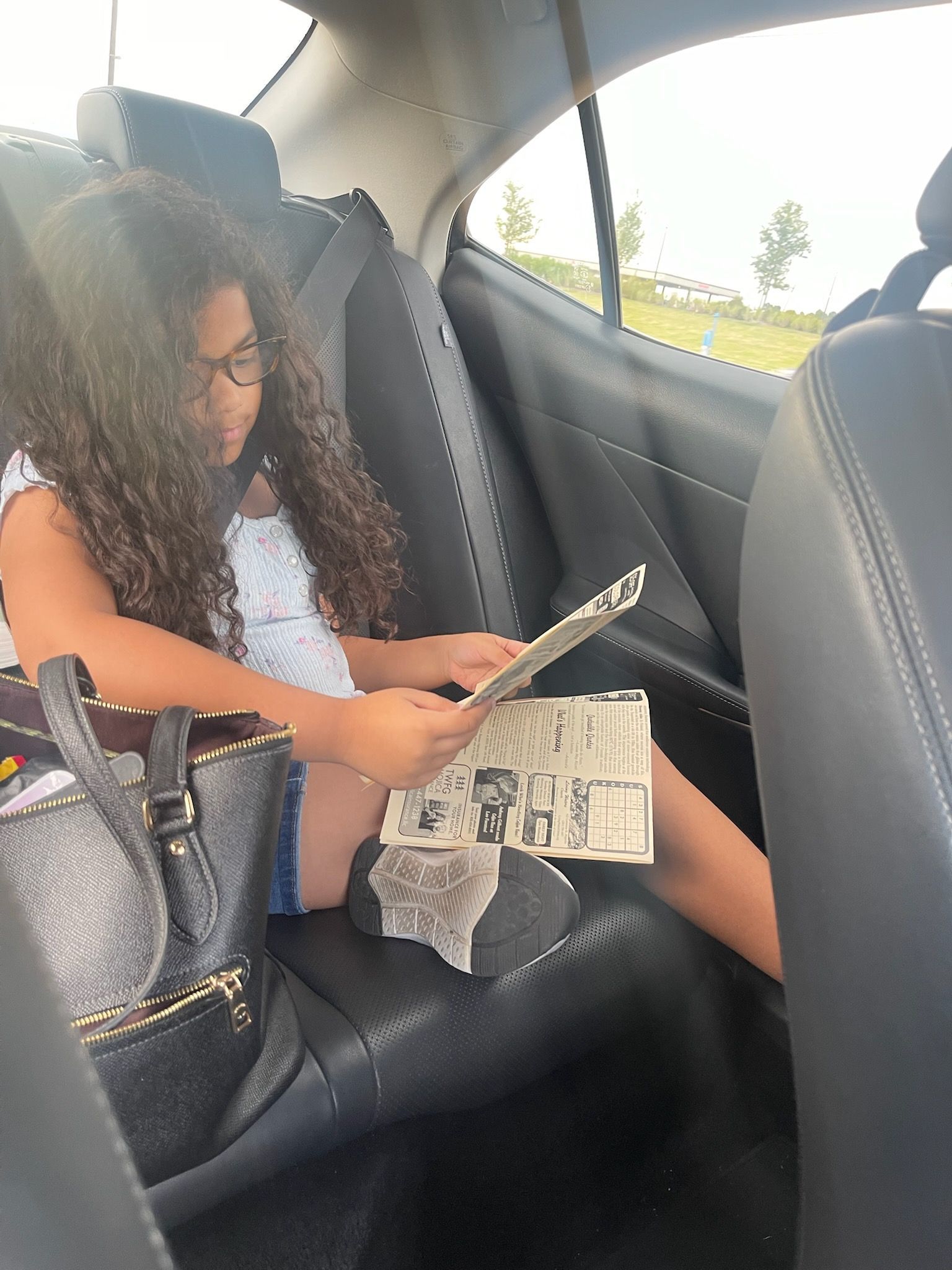 Girl reading newspaper in the backseat of a car, near a purse.