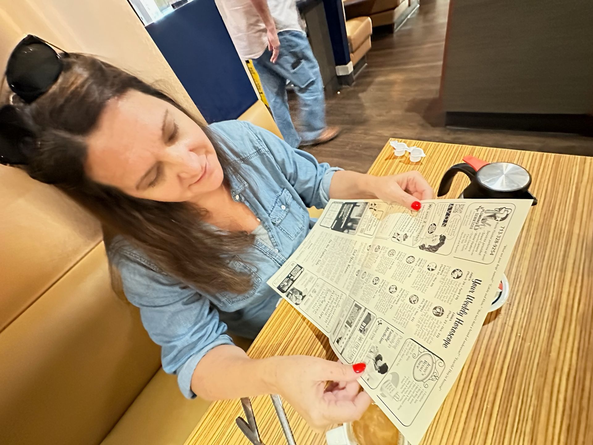 Woman in a denim shirt looking at a newspaper-style menu at a table in a restaurant.